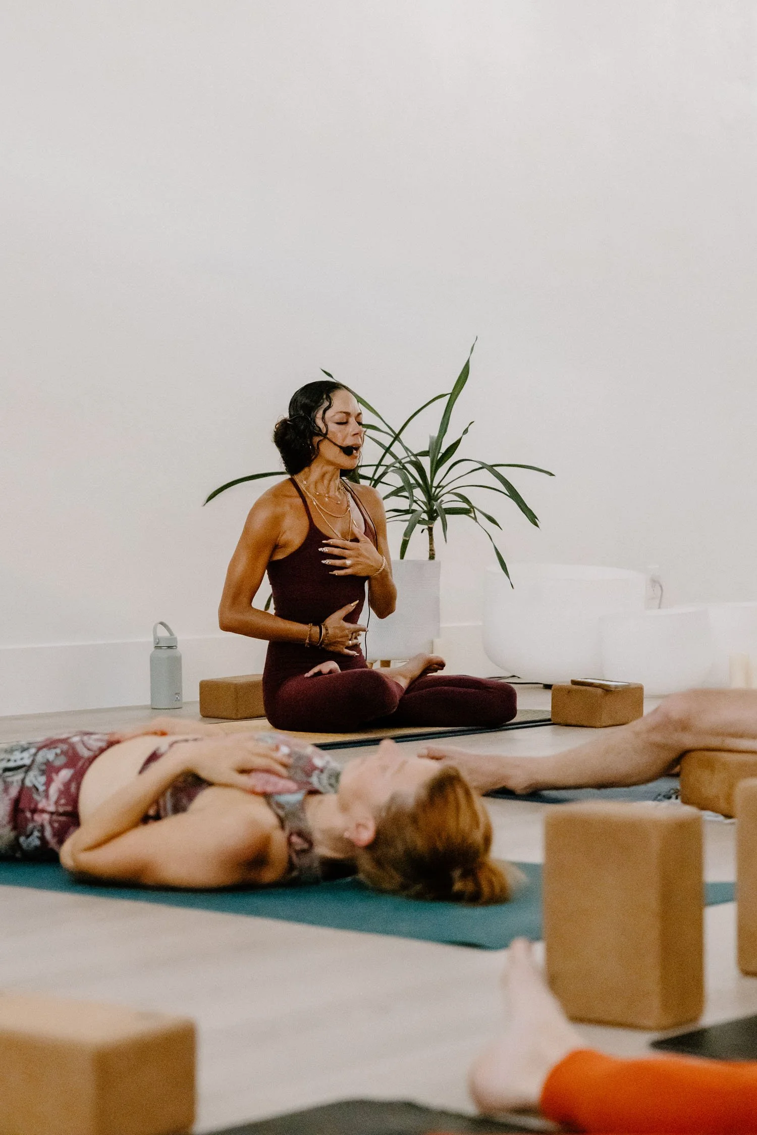 A woman practicing yoga or meditation in a yoga studio, with her eyes closed and hand on her chest, while lying on a yoga mat. Another person is lying on a mat in front of her, with just their legs and part of their body visible. The room has a minimalist decor with a plant and yoga props.