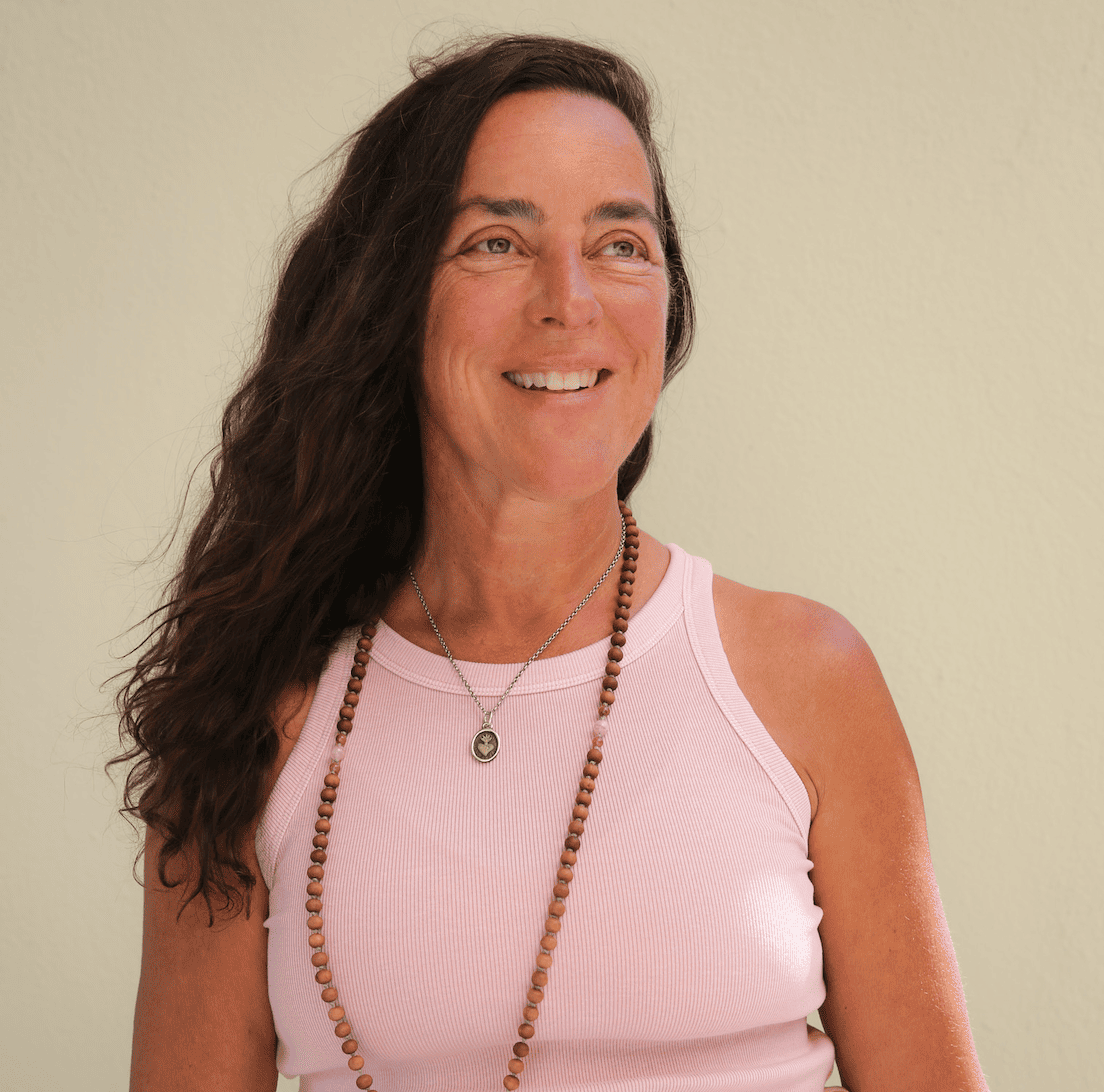 A woman with long wavy brown hair wearing a sleeveless gray top and a gold necklace with a small red pendant, smiling slightly against a plain light-colored background.