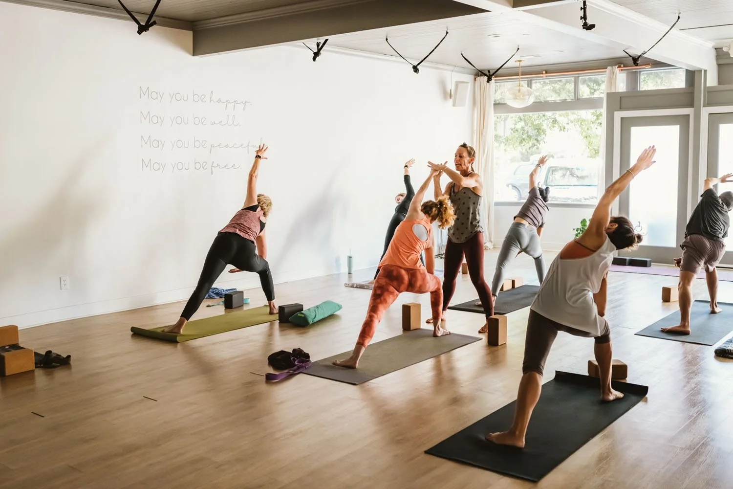 Group of people practicing yoga in a bright studio with large windows and wooden floors, with some performing side stretches and others in different poses.