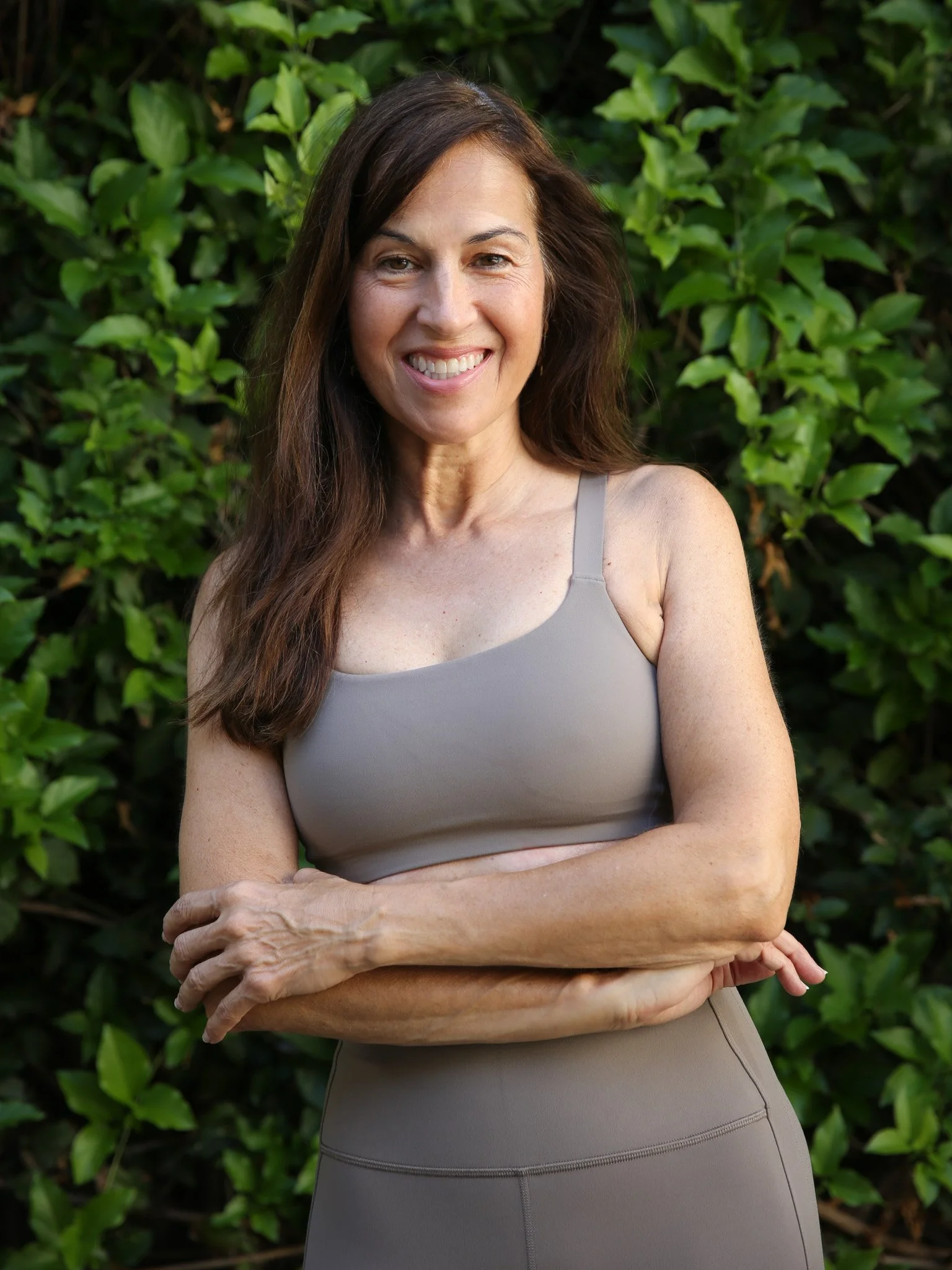 A woman with long brown hair wearing a purple tank top stands outdoors in a forest, smiling and touching a large tree trunk.