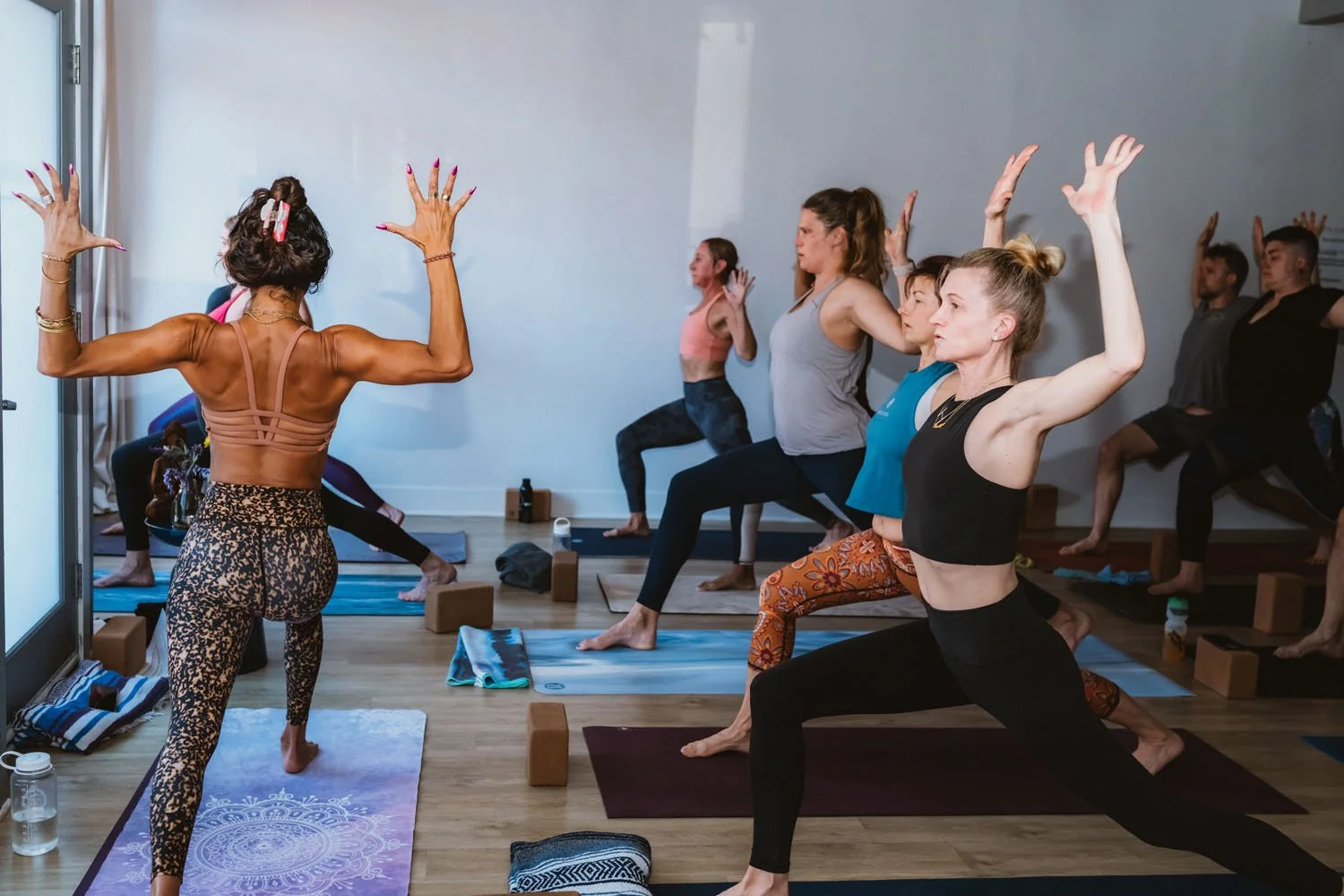 A yoga class with multiple women practicing yoga poses on mats, led by an instructor demonstrating a pose with her arms raised, in a bright room with wooden floors.