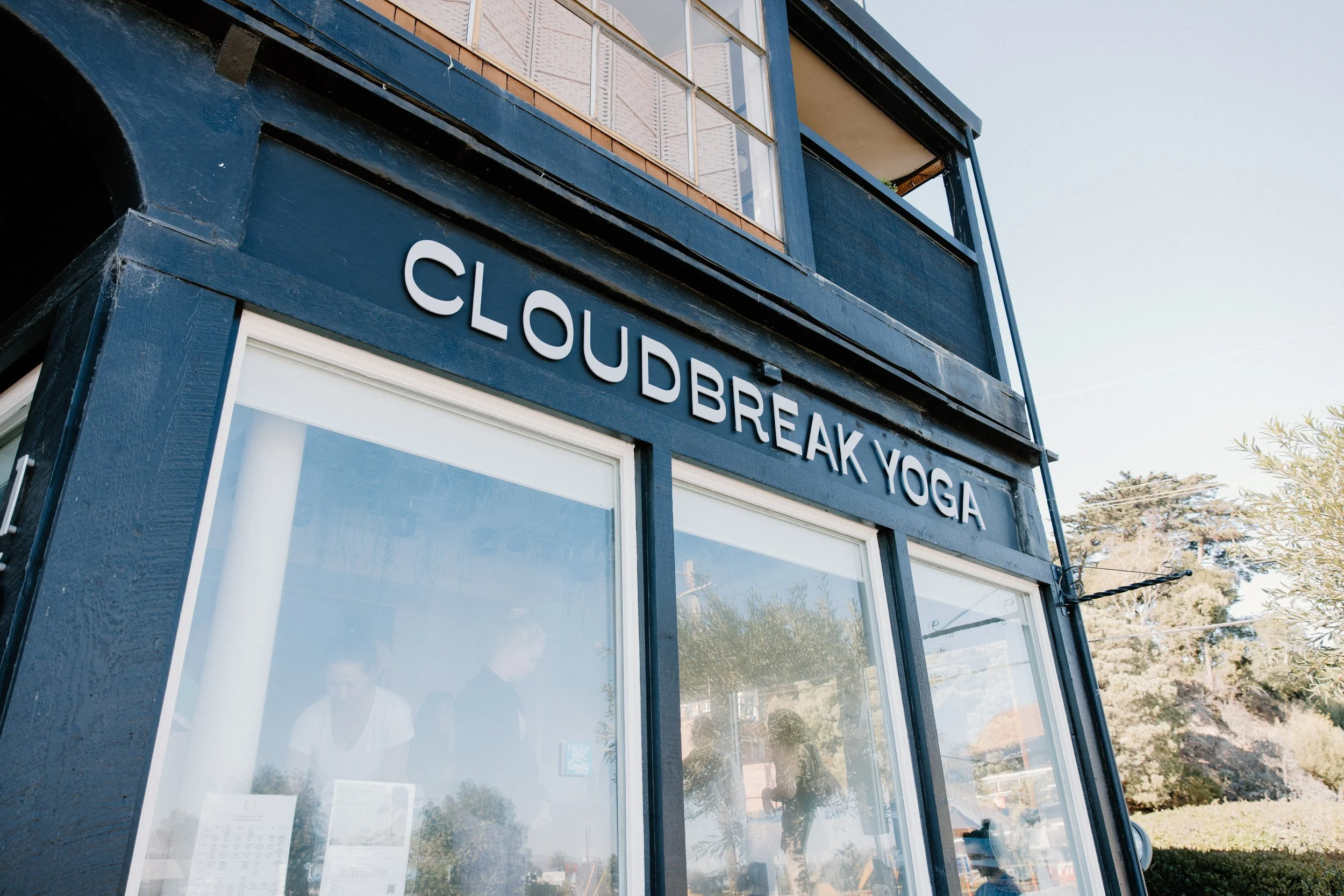 Exterior of a building with a sign that reads 'CLOUDBREAK YOGA' in white letters on a dark background, large windows below the sign showing the interior and reflection of the outside environment.