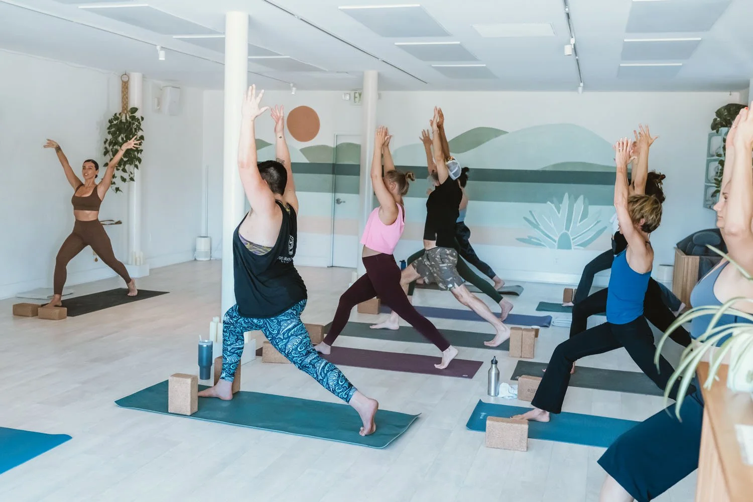 A yoga instructor leading a class of women practicing yoga in a studio, with some in warrior pose, on yoga mats with blocks and water bottles visible.