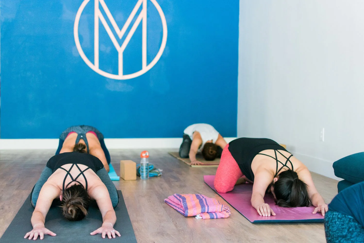 Three women participating in a yoga class, practicing downward dog pose in a room with a blue wall and wooden floor.