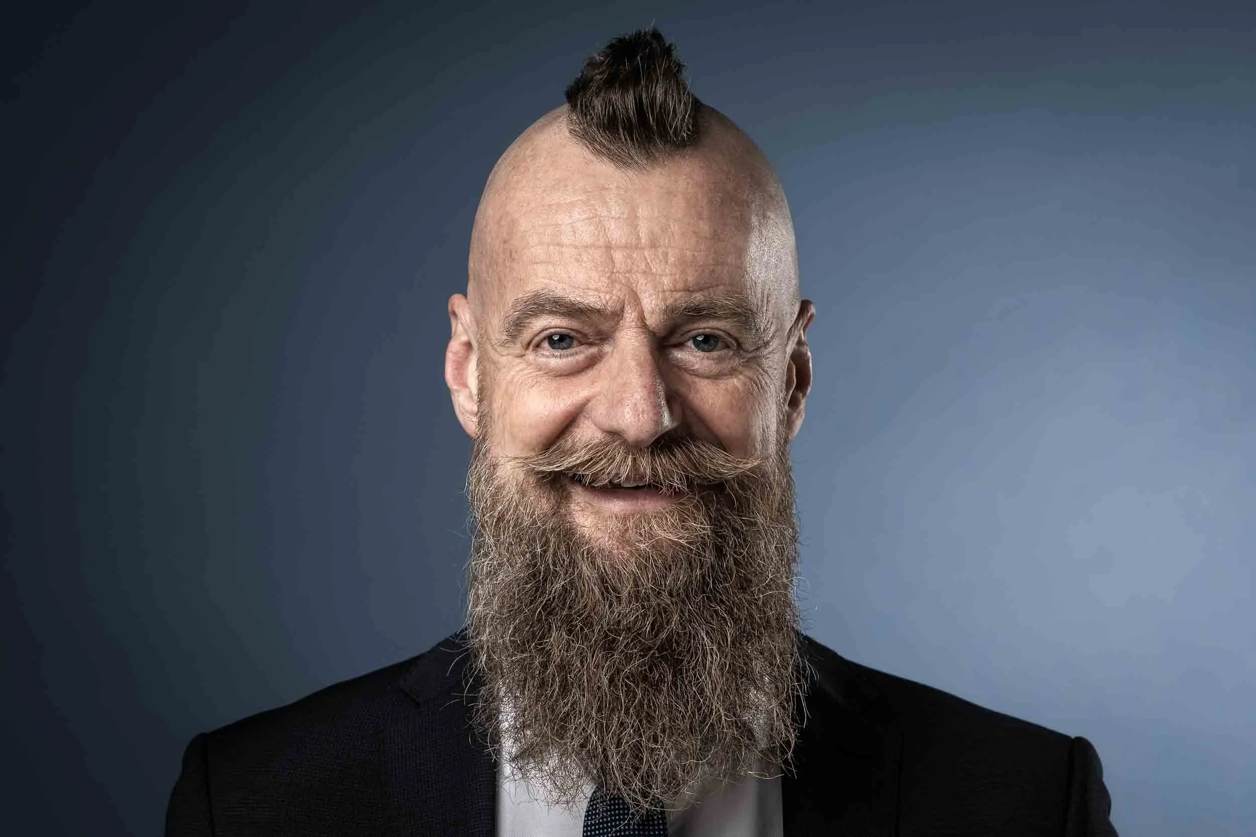 Ein freundlicher Mann mit Glatze, langem Bart, attraktiver Kleidung und einem modernen Haarschnitt, lächelnd vor einem dunklen Hintergrund.