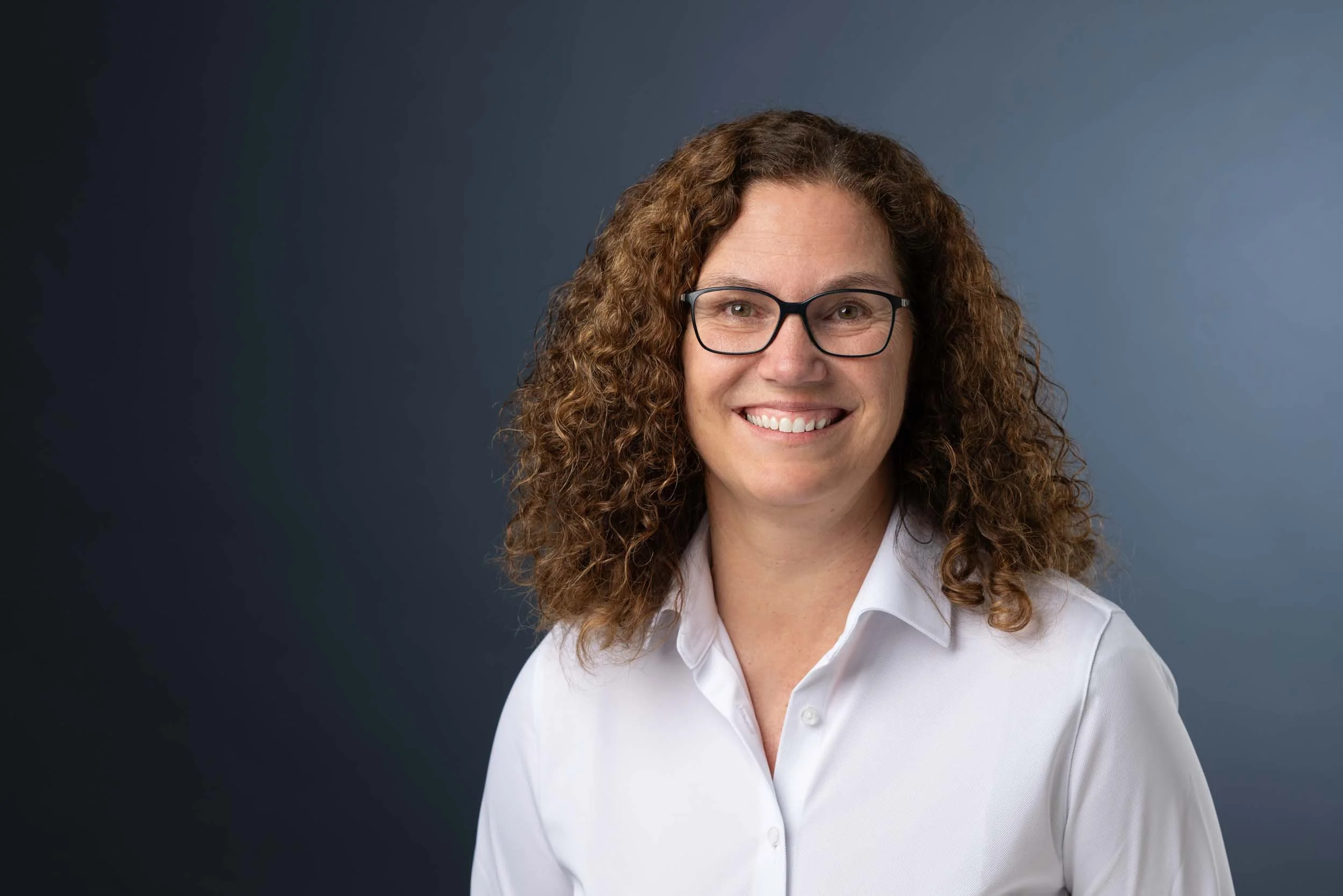 Porträt einer lächelnden Frau mit Brille und lockigem Haar, trägt eine weiße Bluse, vor dunklem Blau-Hintergrund.