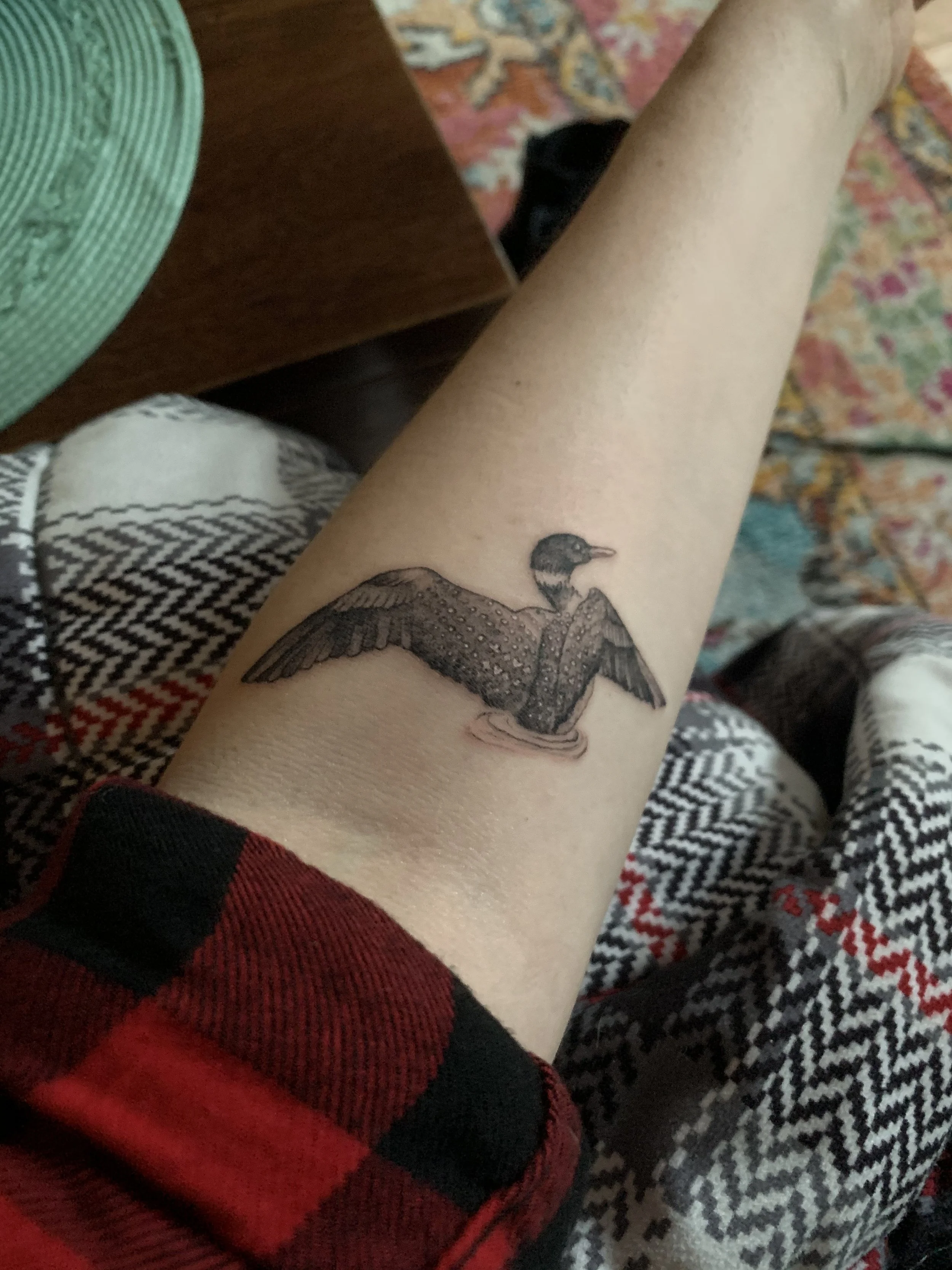 A person's arm with a tattoo of a bird in flight on the forearm, wearing a red and black shirt, with other clothing and a patterned carpet in the background.