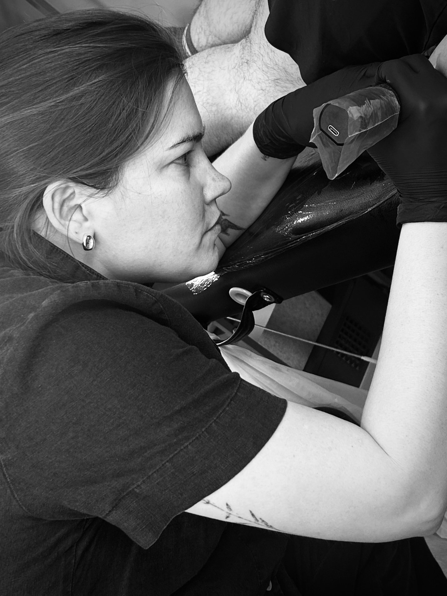 A young woman with light skin and long hair lying back with her head tilted, receiving an injection in her upper arm from a medical professional wearing black gloves. The scene is in a medical setting.