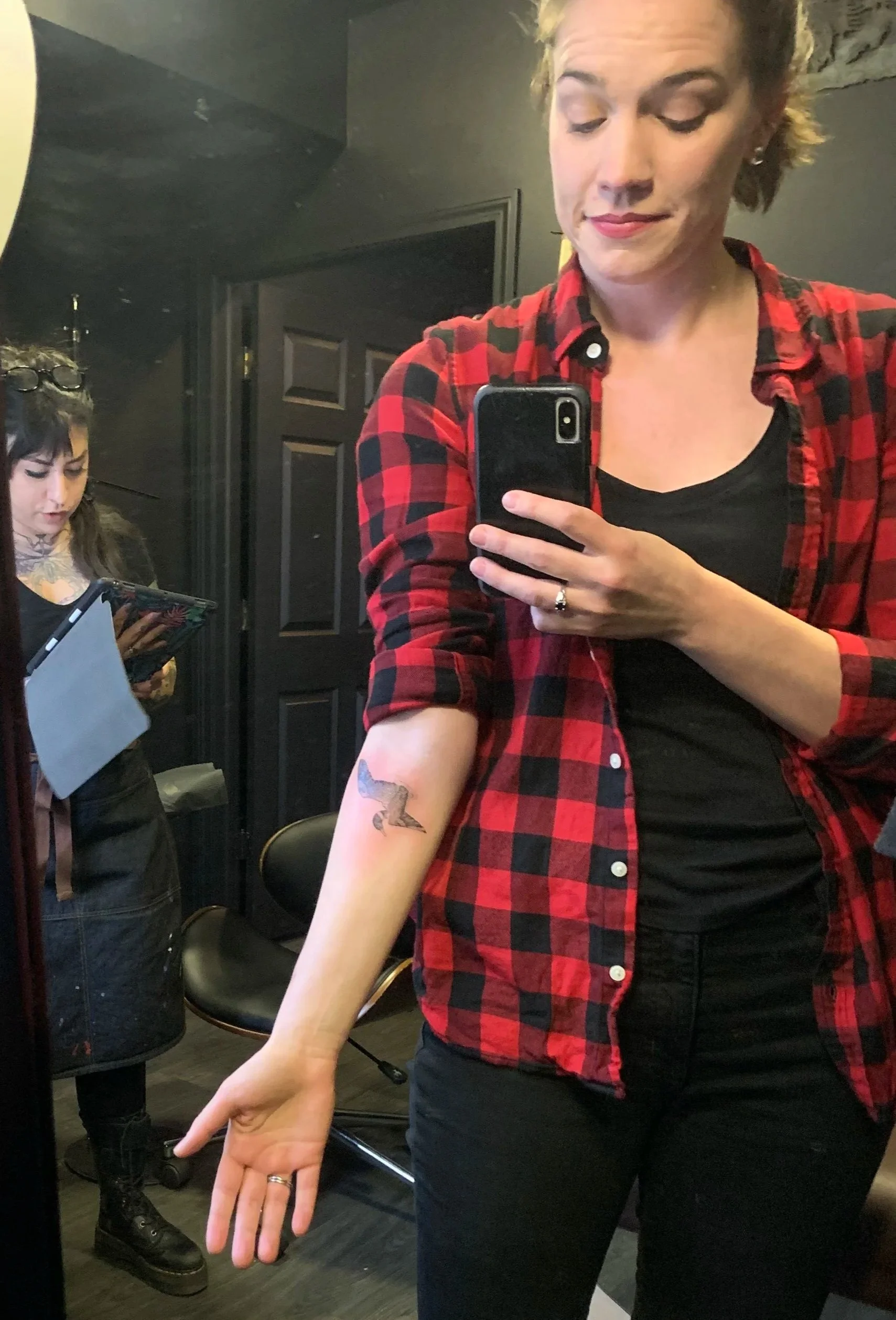 A woman in a red and black checkered shirt smiling while taking a selfie in a mirror. She has short blonde hair, a tattoo of a bird on her forearm, and is wearing a ring on her finger. In the background, there is another woman with glasses on her head, looking at her phone.
