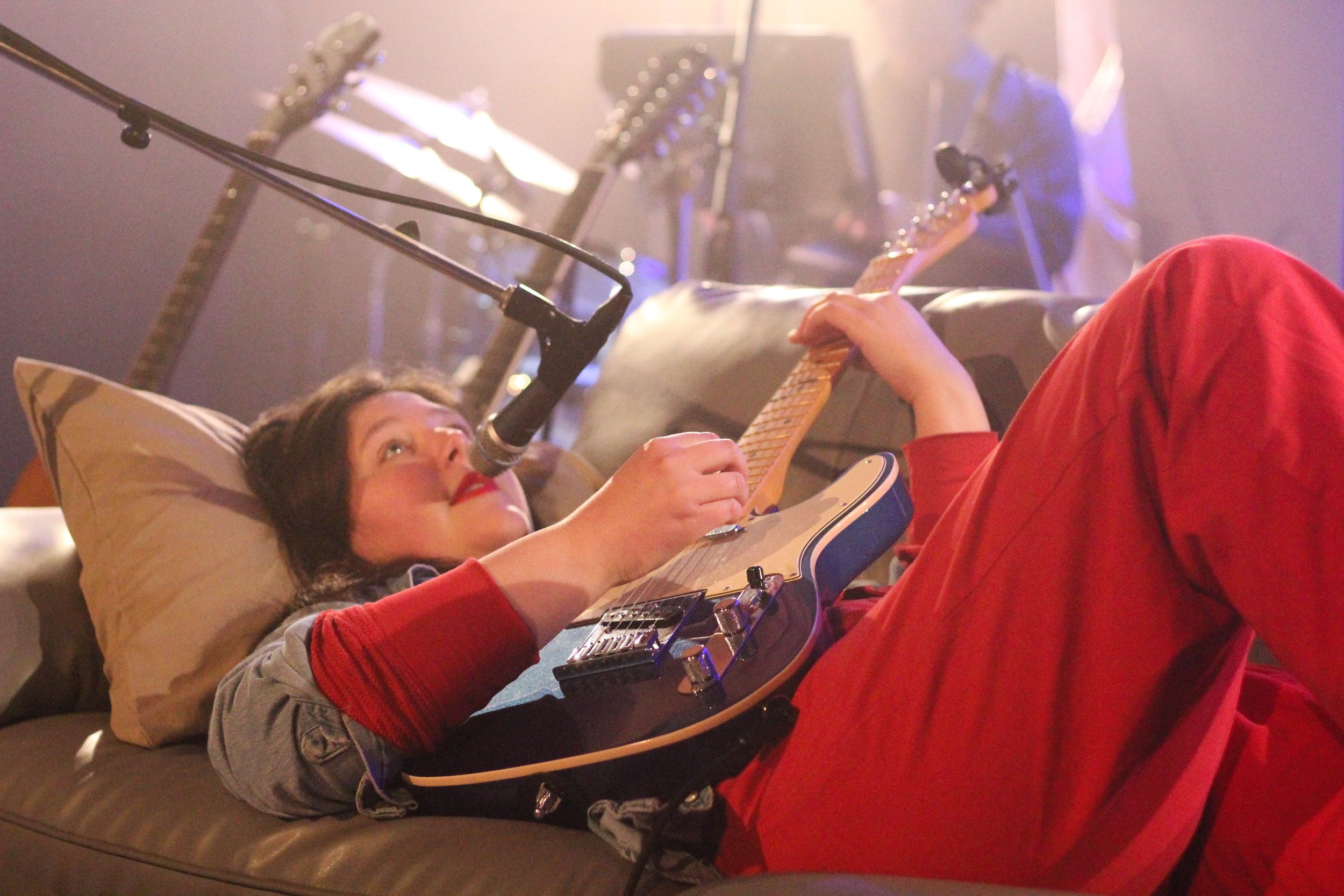 Portrait of Lucy Dacus lying on a couch, playing a blue electric guitar. She is wearing a red jumpsuit and a denim jacket. There is a microphone stand that holds a microphone over her mouth.