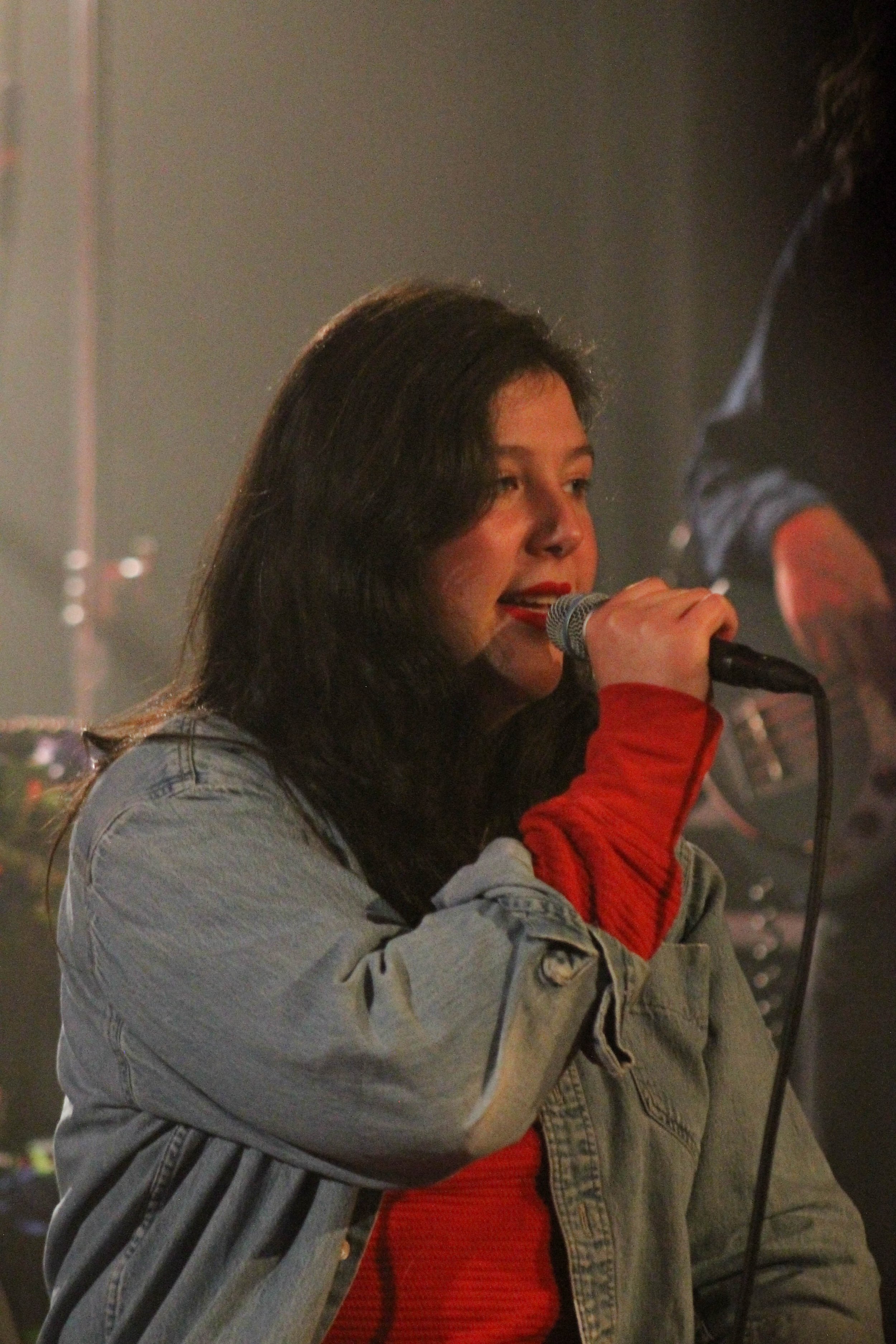 Portrait of Lucy Dacus from the waist up. She is wearing a red jumpsuit and a denim jacket. Her long, dark hair slightly covers one eye. She is holding a microphone in one hand. In the background one arm and an electric guitar are visible.