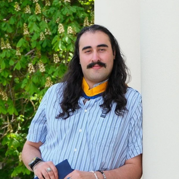 Luca Morones-Calcagno, a young man with long, dark curly hair smiling slightly in front of a background of green plants and a white wall.