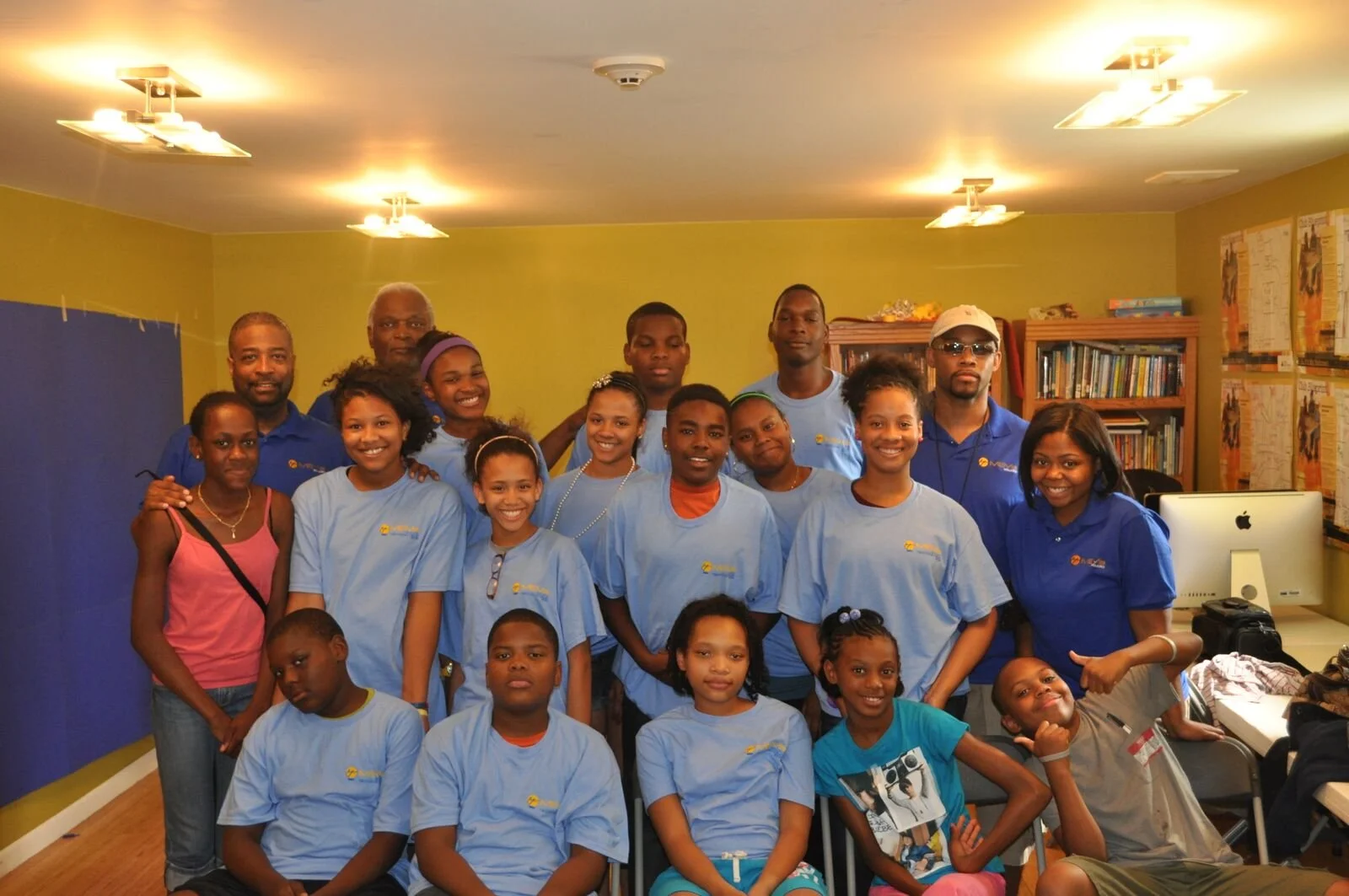 Group of children and adults posing together in a room, some wearing matching blue shirts, indicating a team or organization event.