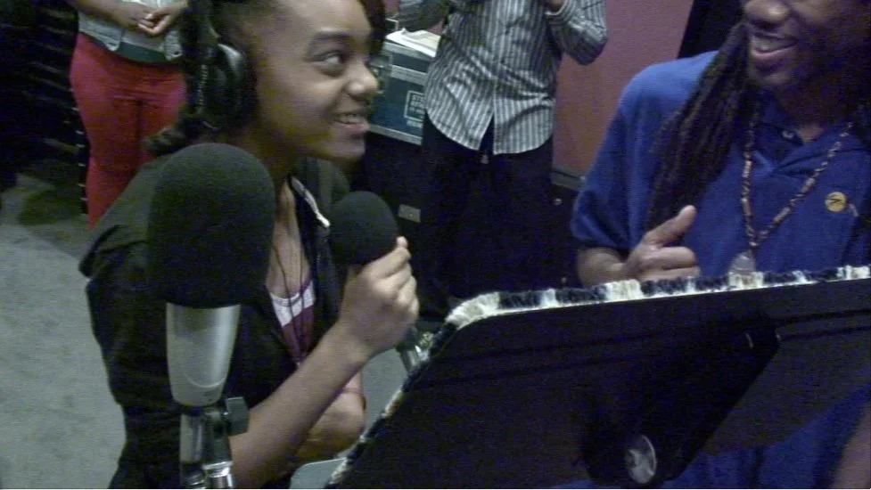 A young girl is wearing headphones and singing into a microphone at a radio station or recording studio, with a man standing beside her, smiling and giving a thumbs-up gesture.