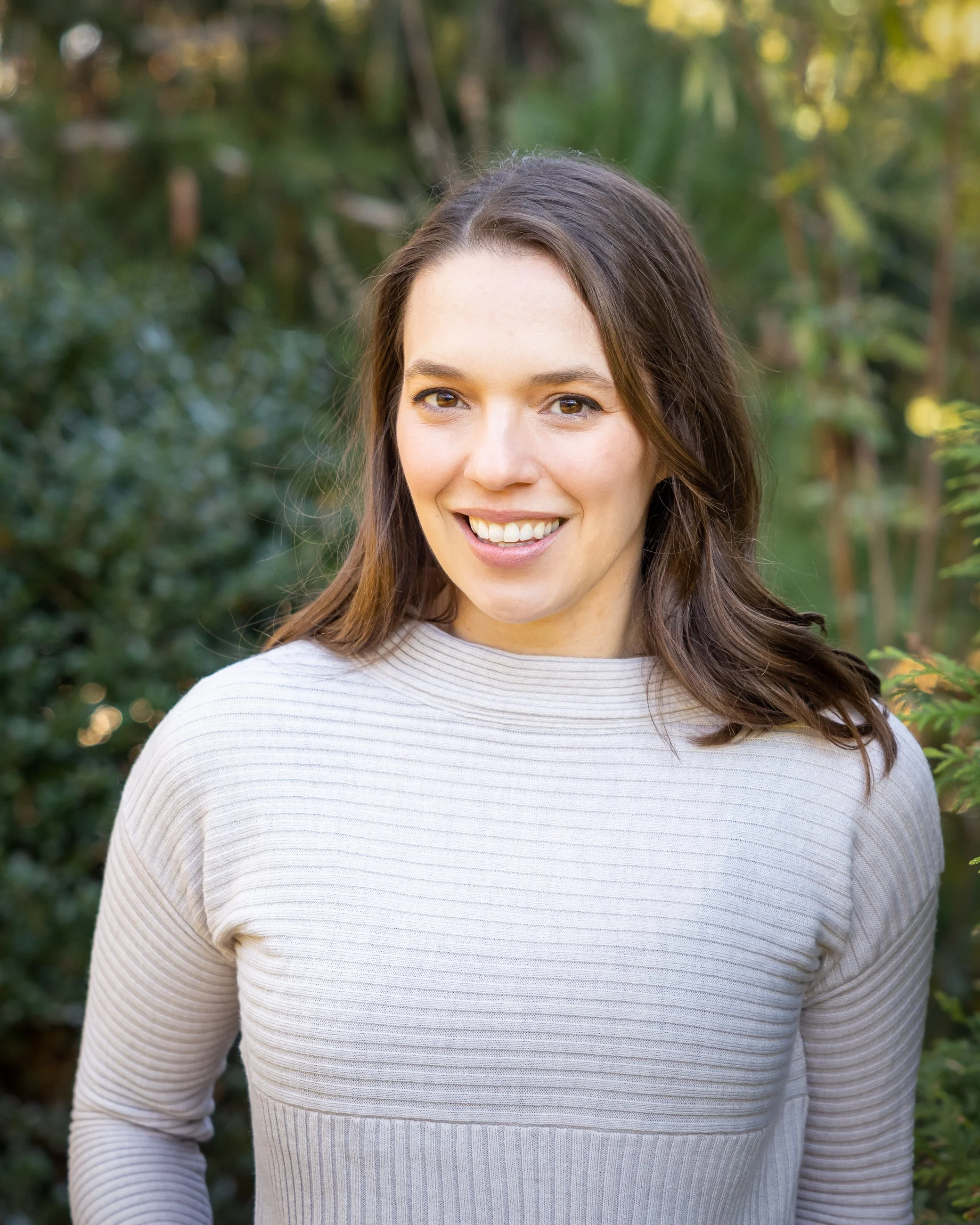 Dr. Stephanie Ernestus is licensed child psychologist and therapist who works with kids, teens, and families. She is pictured here standing in front of greenery wearing a purple sweater.