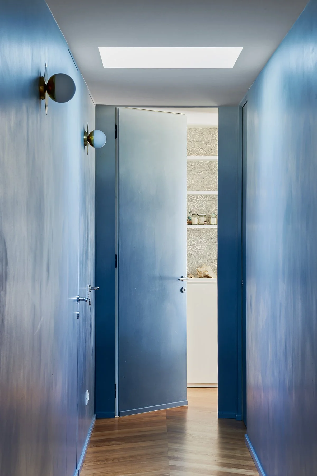 Looking down a deep blue corridor to a fresh white space beyond, glimpsing through to shelves lined with Abigail Edwards' Seascape waves wallpaper