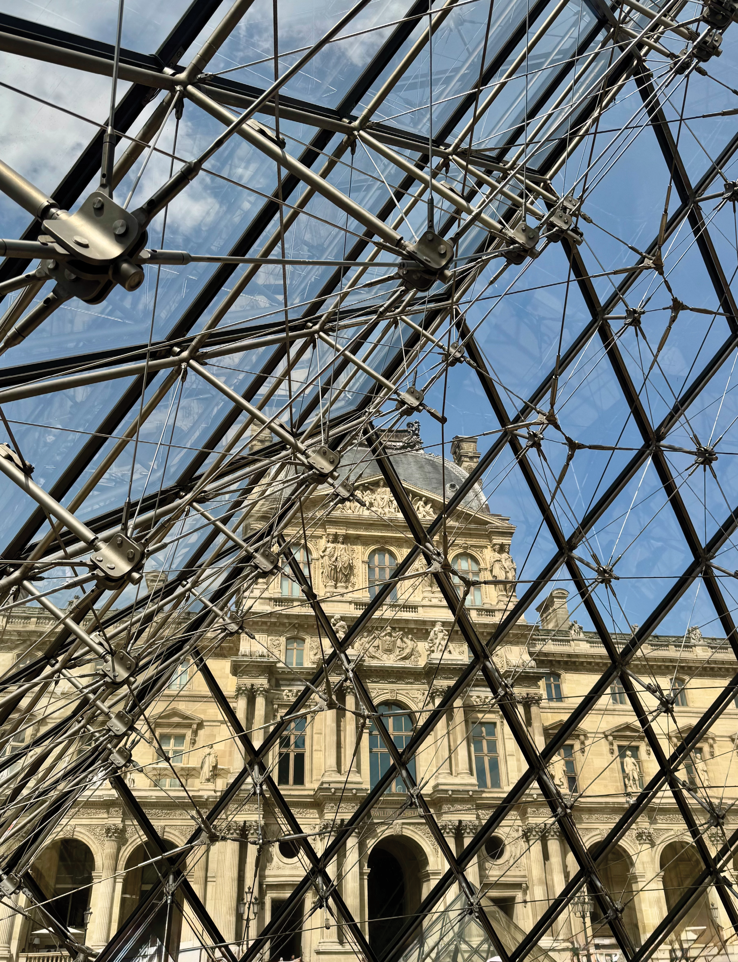 a view from inside the iconic Louvre Pyramid in Paris, looking out toward the historic French Renaissance facade of the Louvre Museum