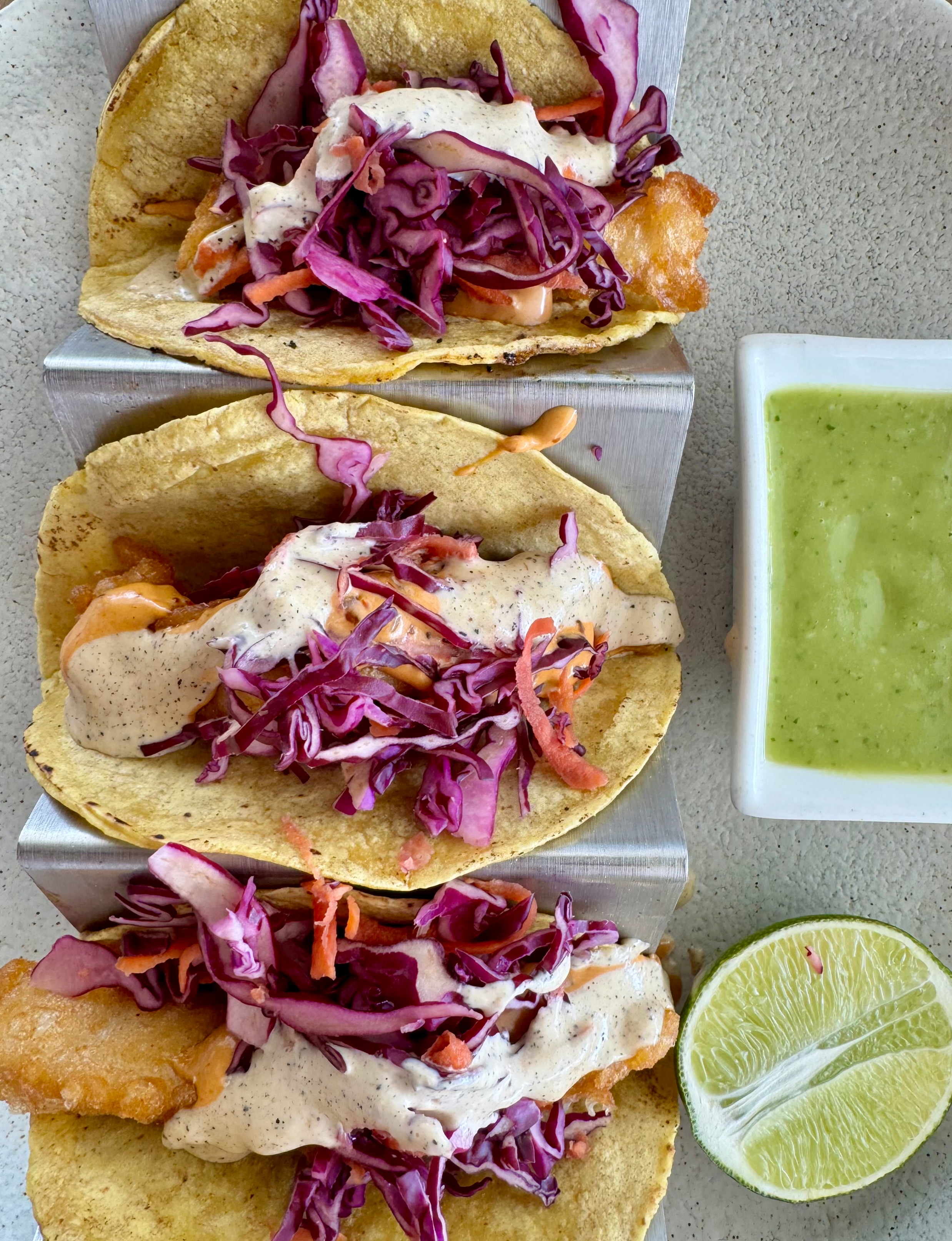 Mexican cuisine three fish tacos topped with shredded purple cabbage and carrots and drizzled with a creamy white sauce and side of bright green salsa and a fresh lime half