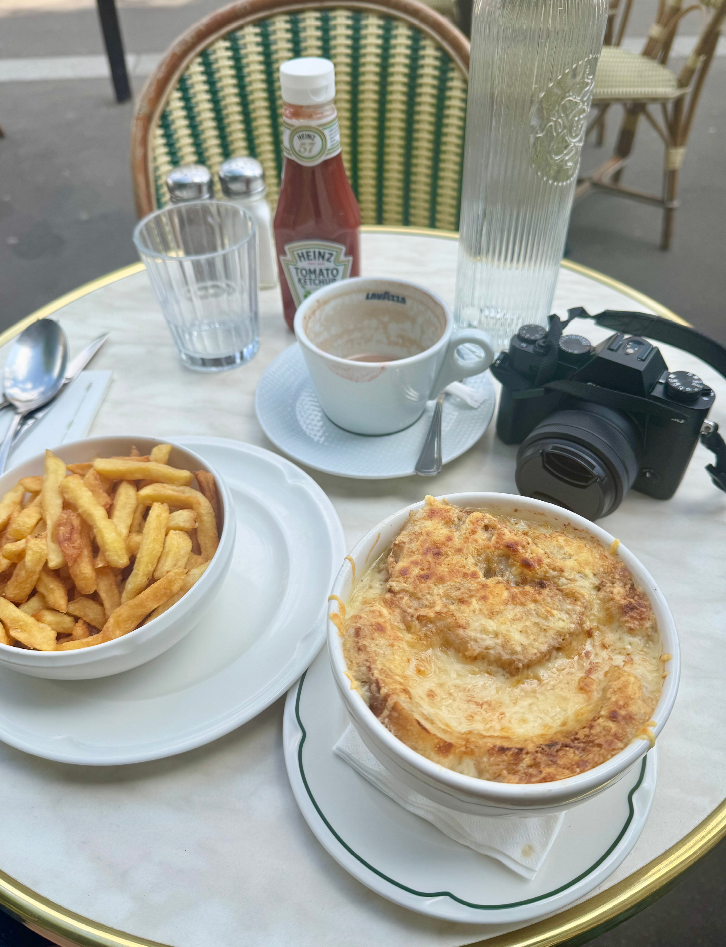 a classic French café table set with a bowl of French onion soup topped with melted cheese, a side of french fries, and a camera