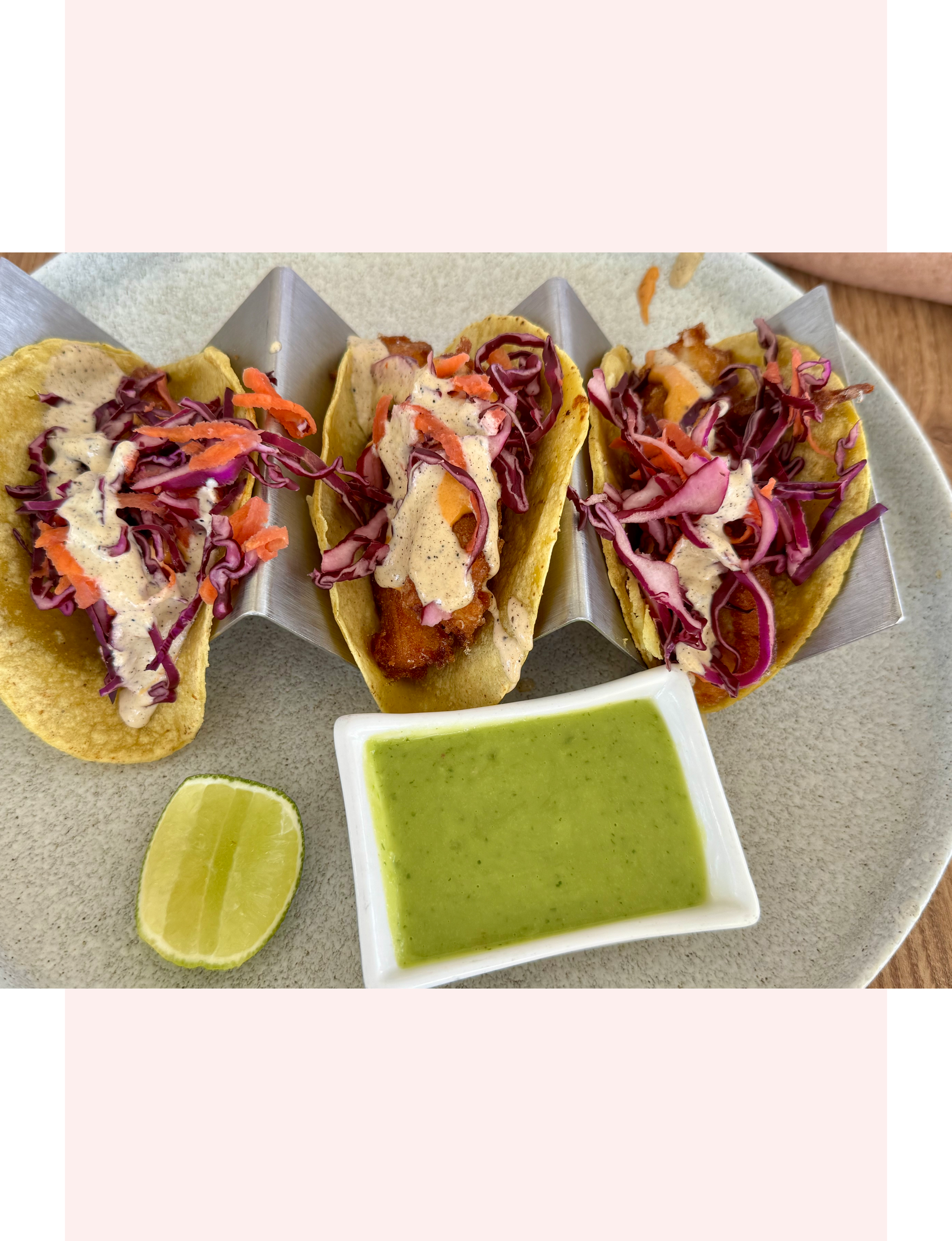 Mexican cuisine three fish tacos topped with shredded purple cabbage and carrots and drizzled with a creamy white sauce and side of bright green salsa and a fresh lime half