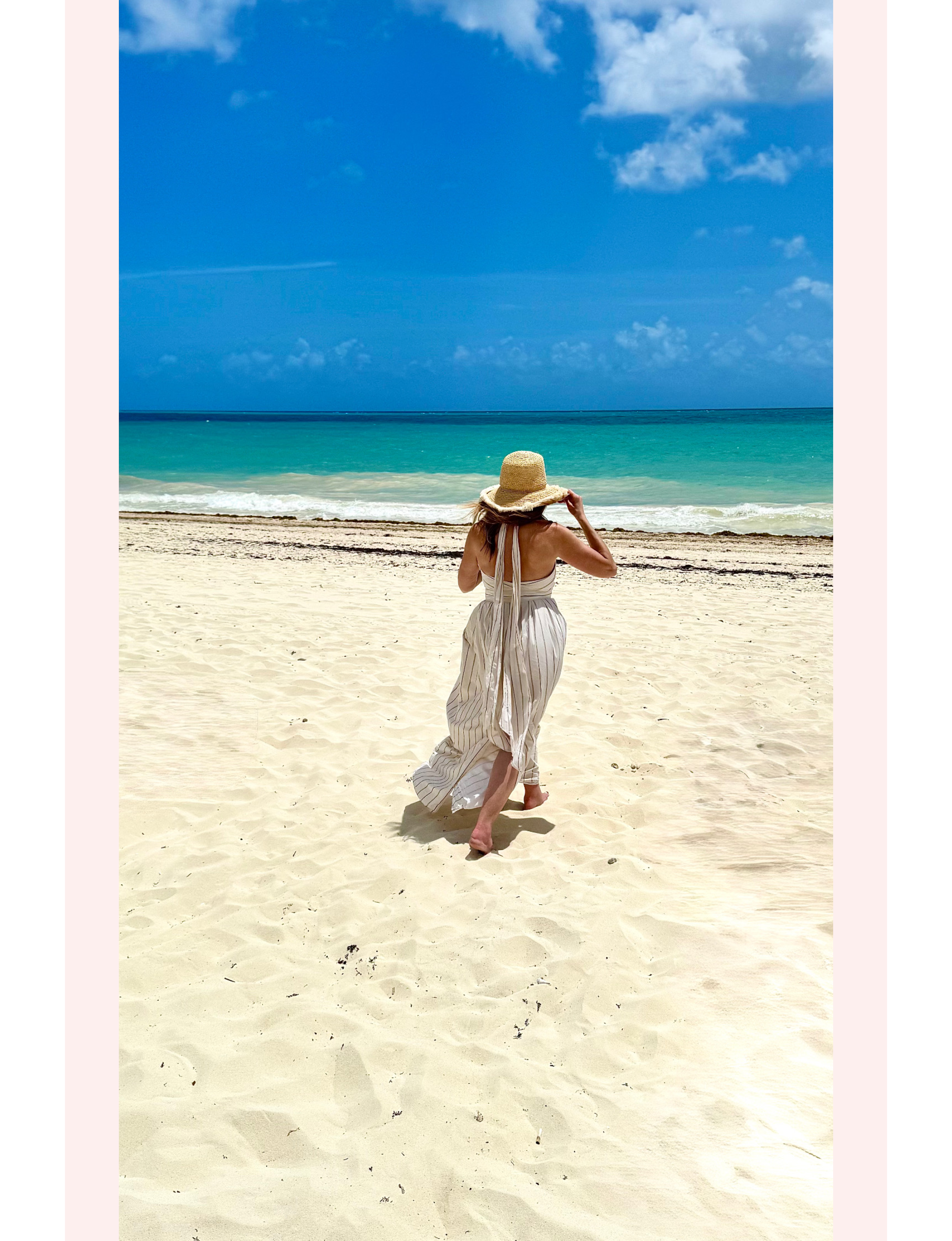 Michele Rosenthal walking along a white-sand tropical beach wearing a striped halter maxi dress and a straw sun hat