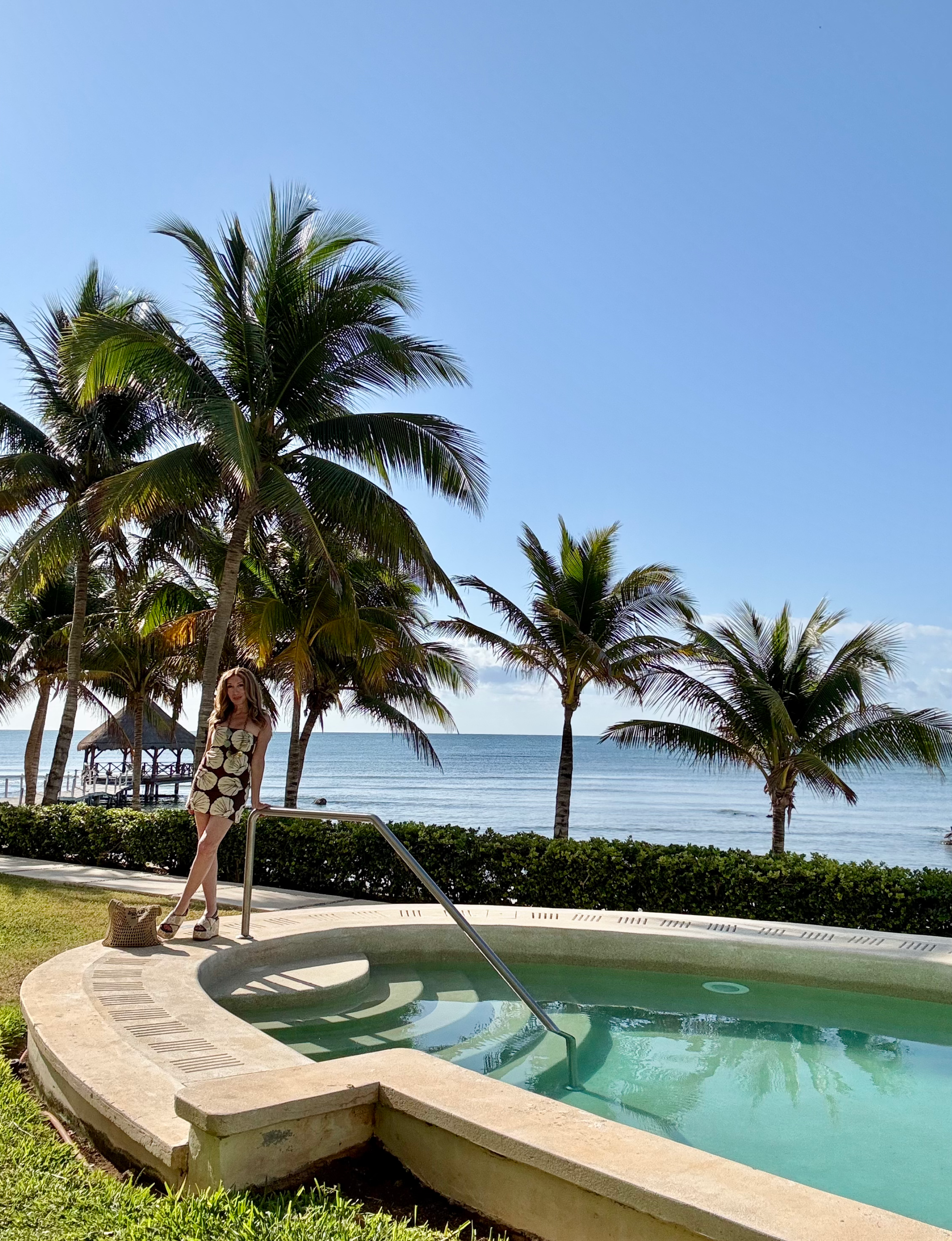 Michele Rosenthal posing by a scenic coastal pool with palm trees and ocean in the background at Riviera Maya in Mexico