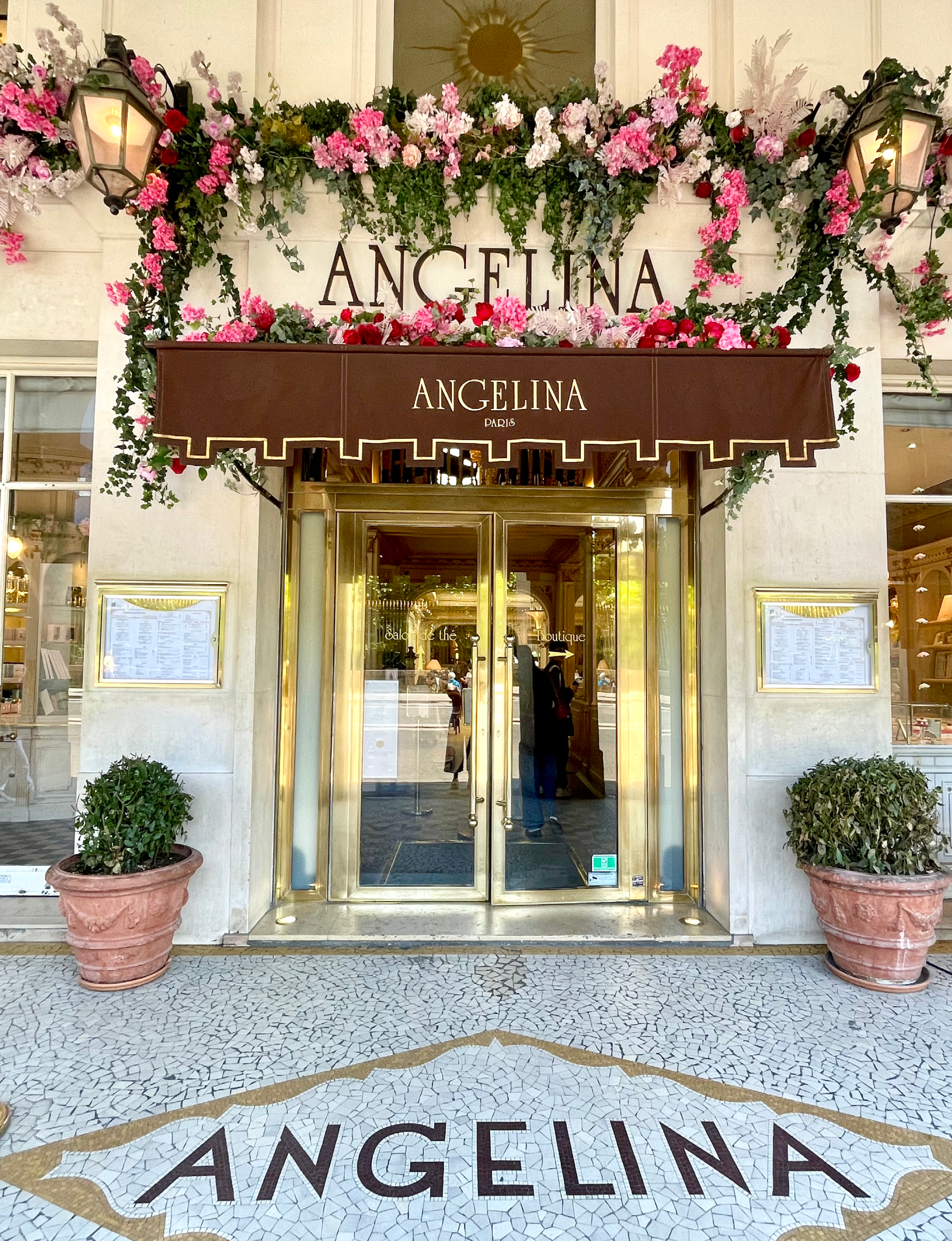 the entrance to the iconic historic tearoom Angelina Paris featuring elegant gold-trimmed double doors, mosaic tile lettering on the ground, and a seasonal floral awning display