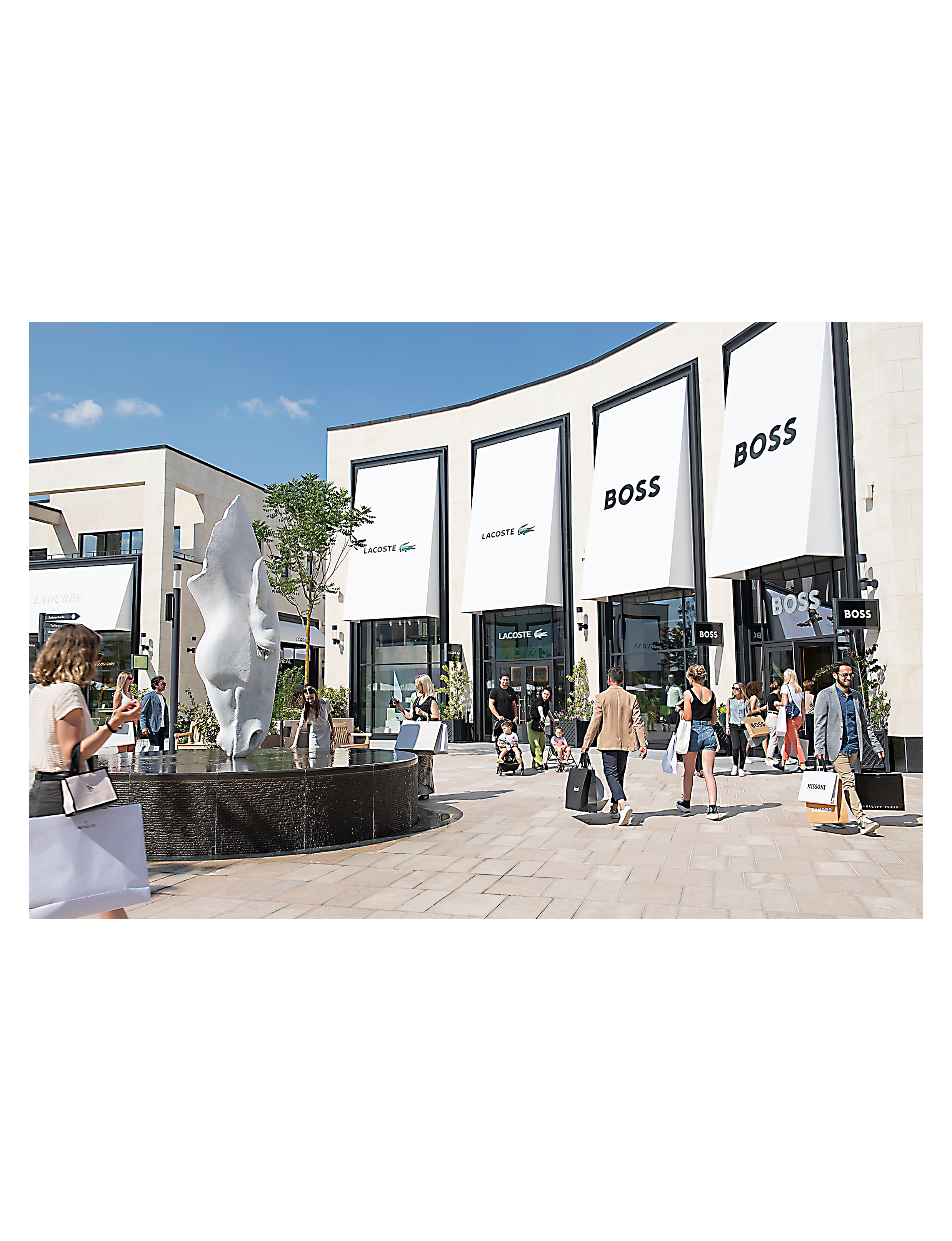 the open-air pedestrian plaza at the McArthurGlen Paris-Giverny Designer Outlet located in Normandy/Giverny region west of Paris, featuring storefronts like BOSS and Lacoste alongside a modern water fountain sculpture
