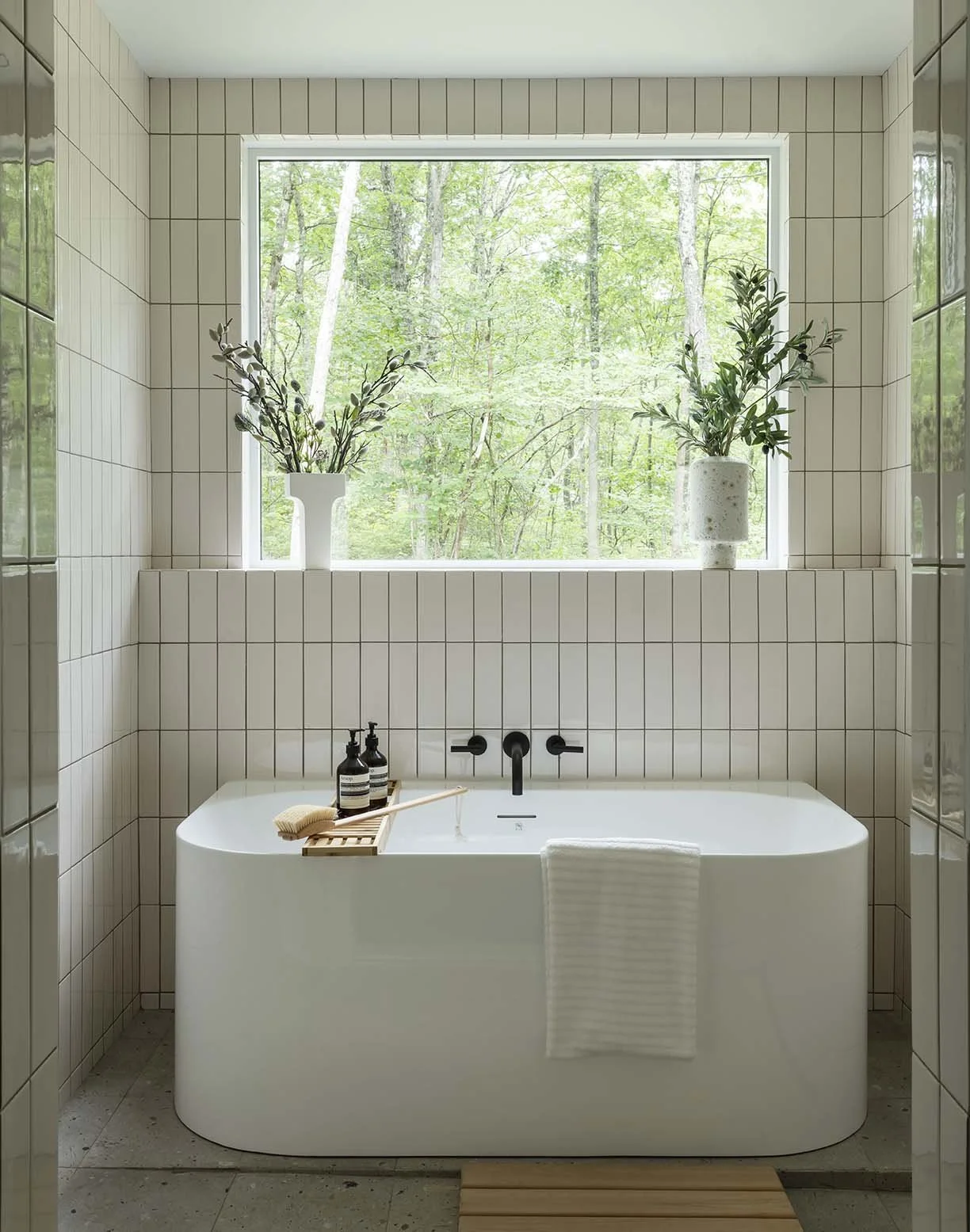 Modern bathroom with a white bathtub, black wall-mounted faucet, and large window with greenery outside. The window sill has two white vases with green branches, and a wooden bath tray with soap, brushes, and bottles.
