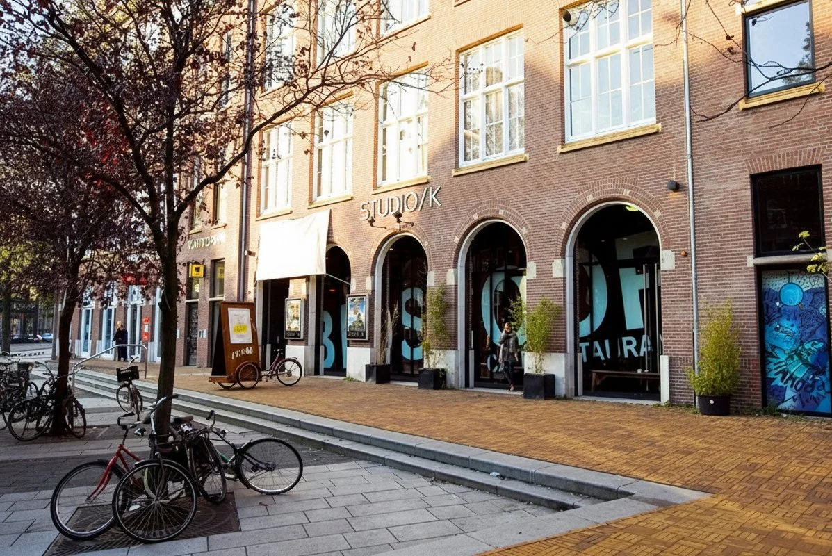 Street view of a brick building with large windows, trees, bicycles, and a sidewalk with pedestrians. The building has a sign that reads 'STUDIO/K' and display windows featuring artwork, with an outdoor chalkboard sign on the sidewalk.