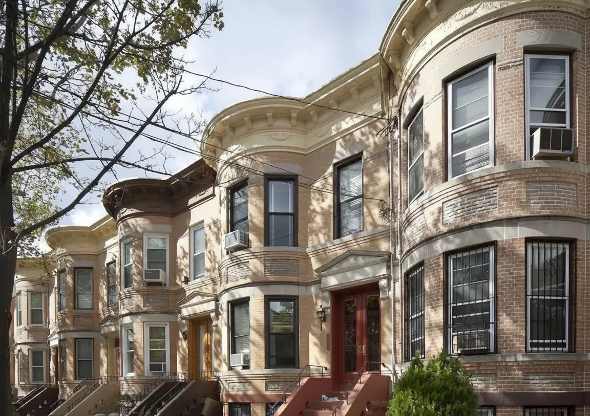 A row of historic brownstone buildings with bay windows, trees, and steps leading to front doors, typical of urban residential neighborhoods.