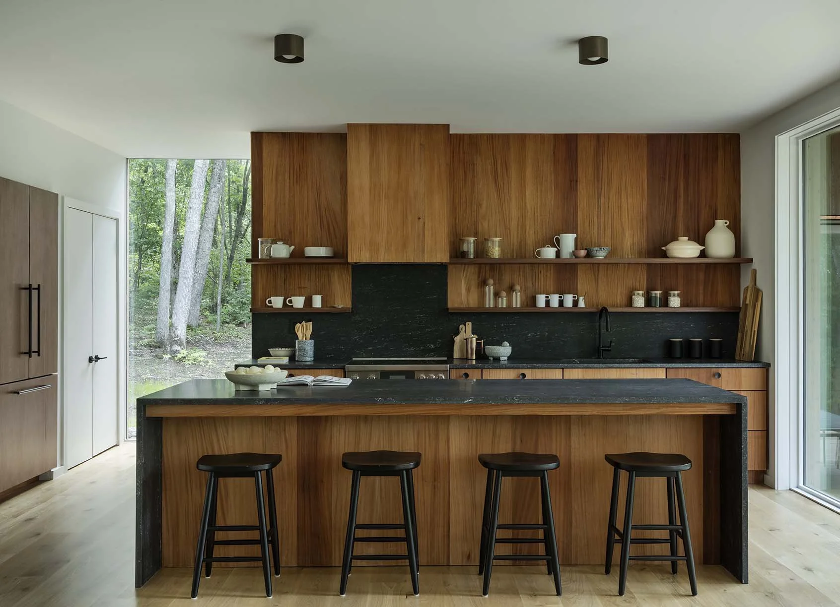 Modern kitchen with wooden cabinets, black countertops, and large windows showing trees outside.