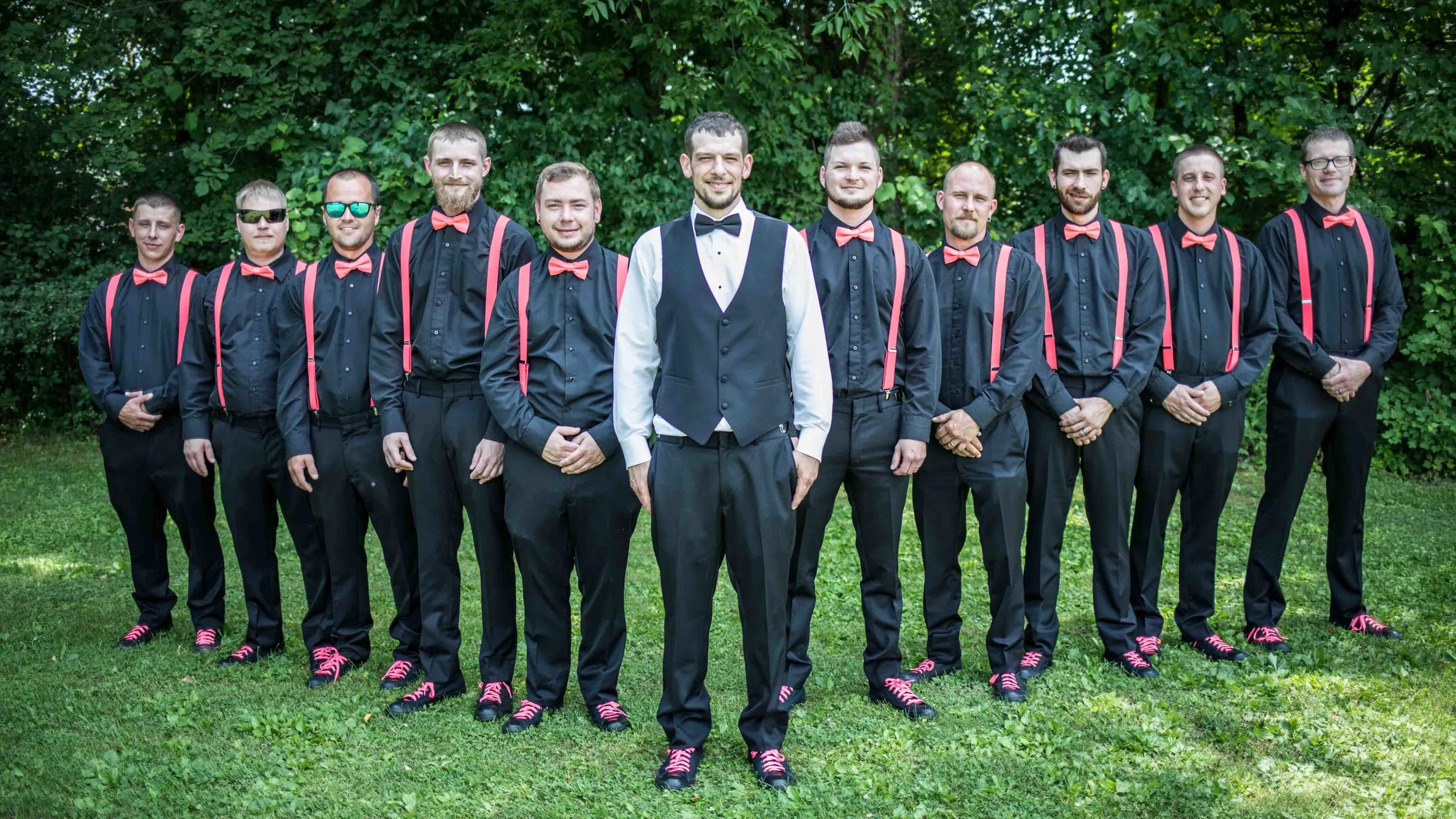 Groom stands up front and center as the 10 other groomsmen wearing black with pink suspenders and bowtie, stand in a v-shape behind him