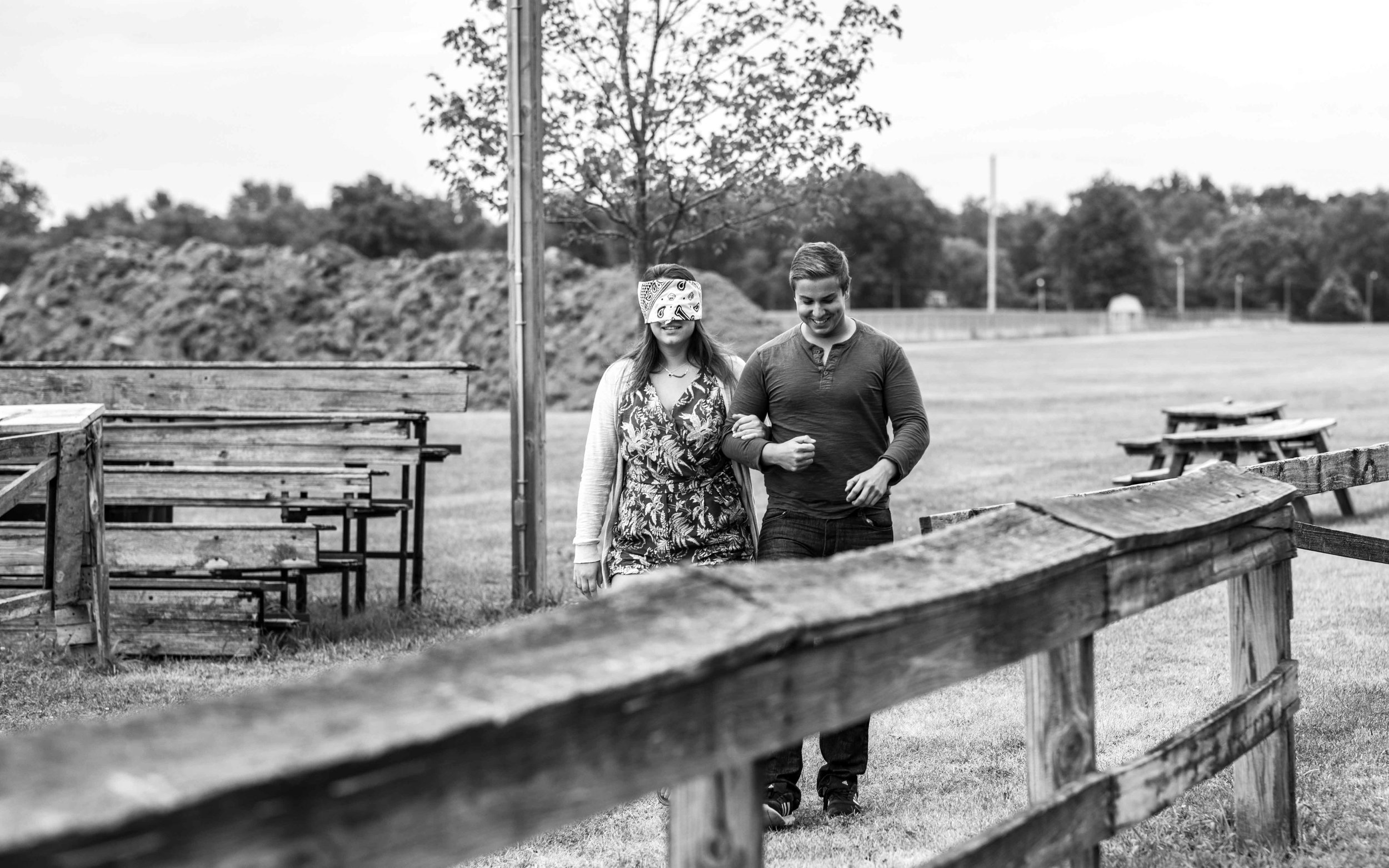  A man leading his blindfolded girlfriend along a split rail fence 