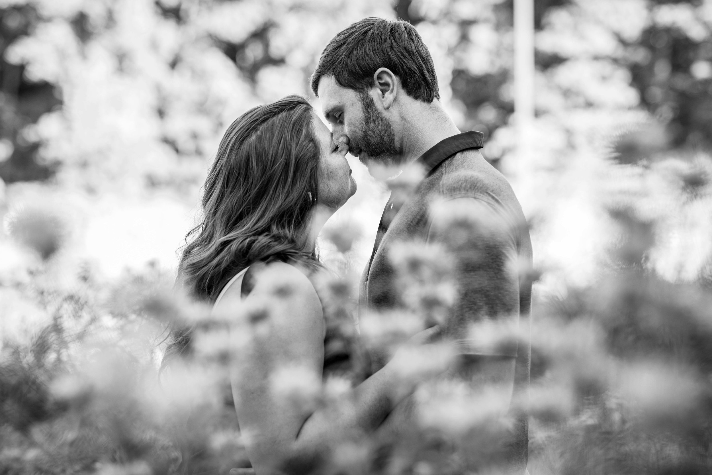  black and white image of a woman kissing her fiancé with wildflowers all around  