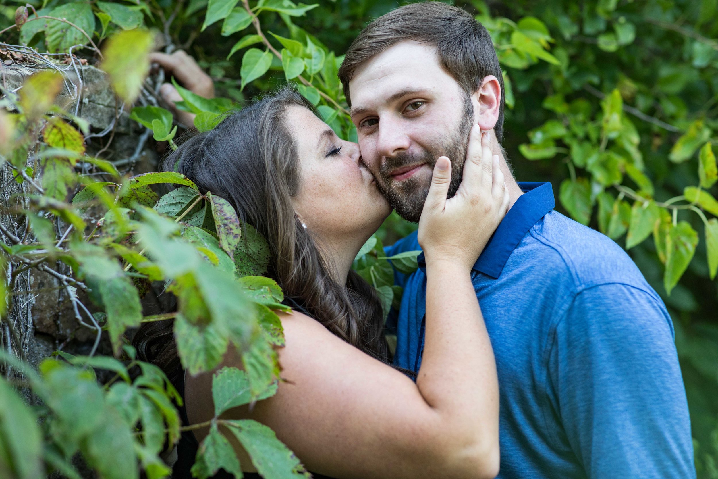  woman kisses a mans cheek as he looks into the camera, they’re both leaning against a stone wall covered in ivy 