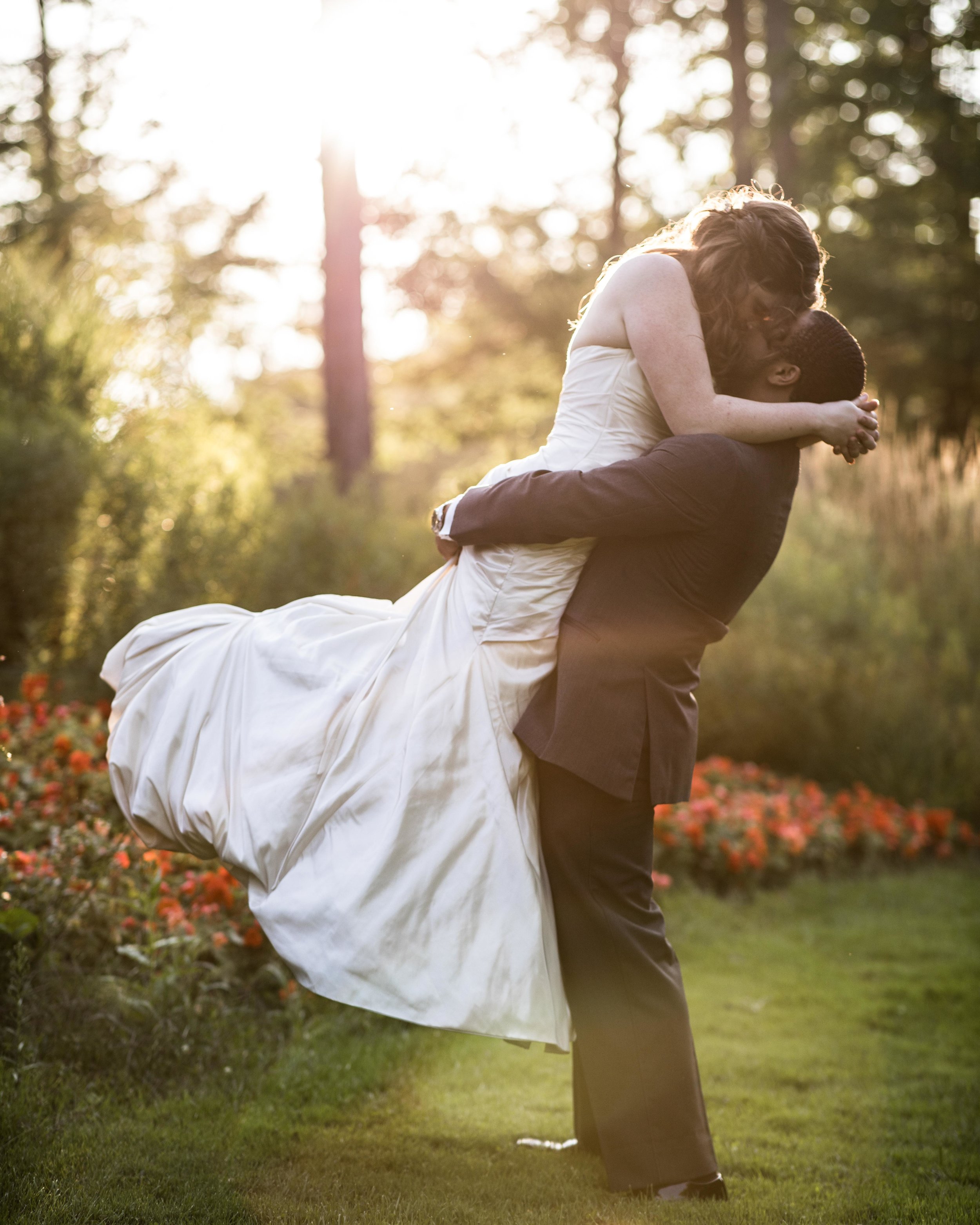  The groom lifts the bride up in the air as she kisses him with sunset and flower landscape behind them 