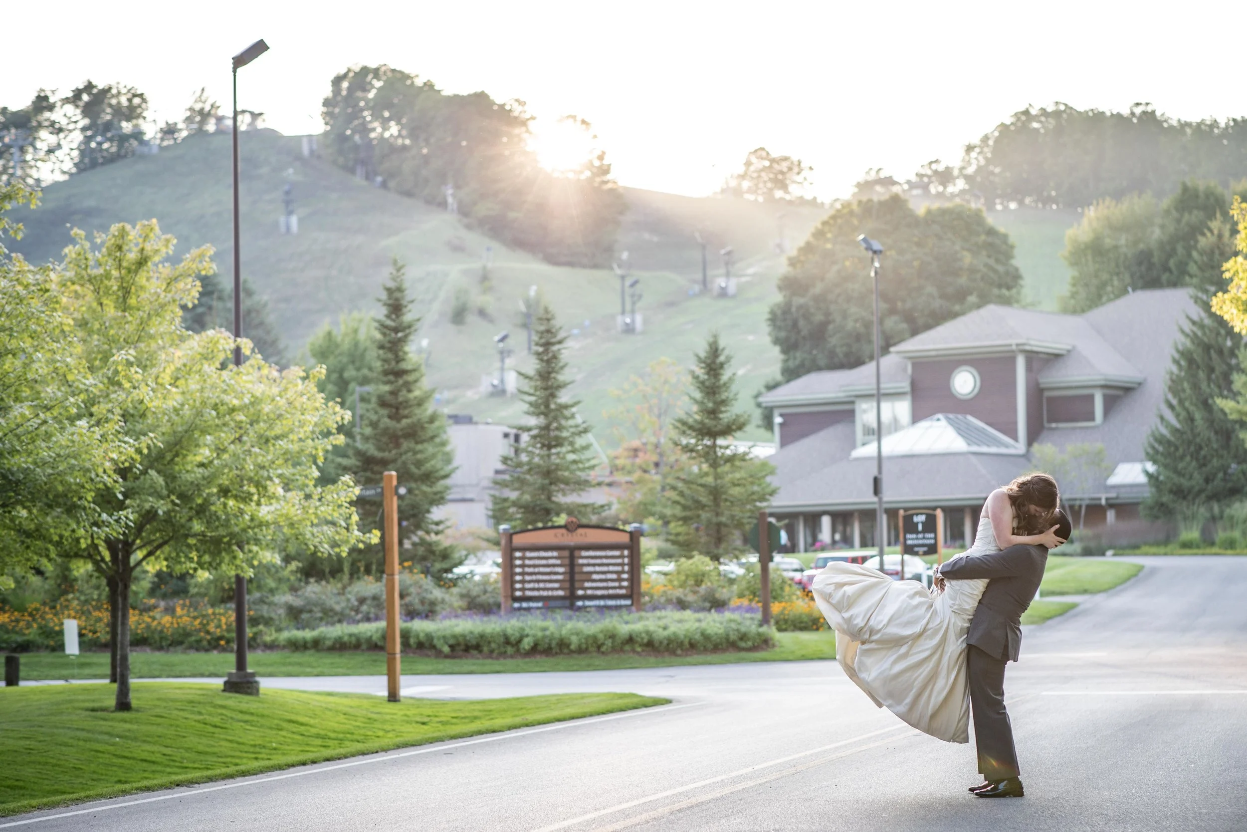 dramatic landscape photo of crystal mountain in Michigan with groom lifting bride as she kisses him during sunset over the mountains