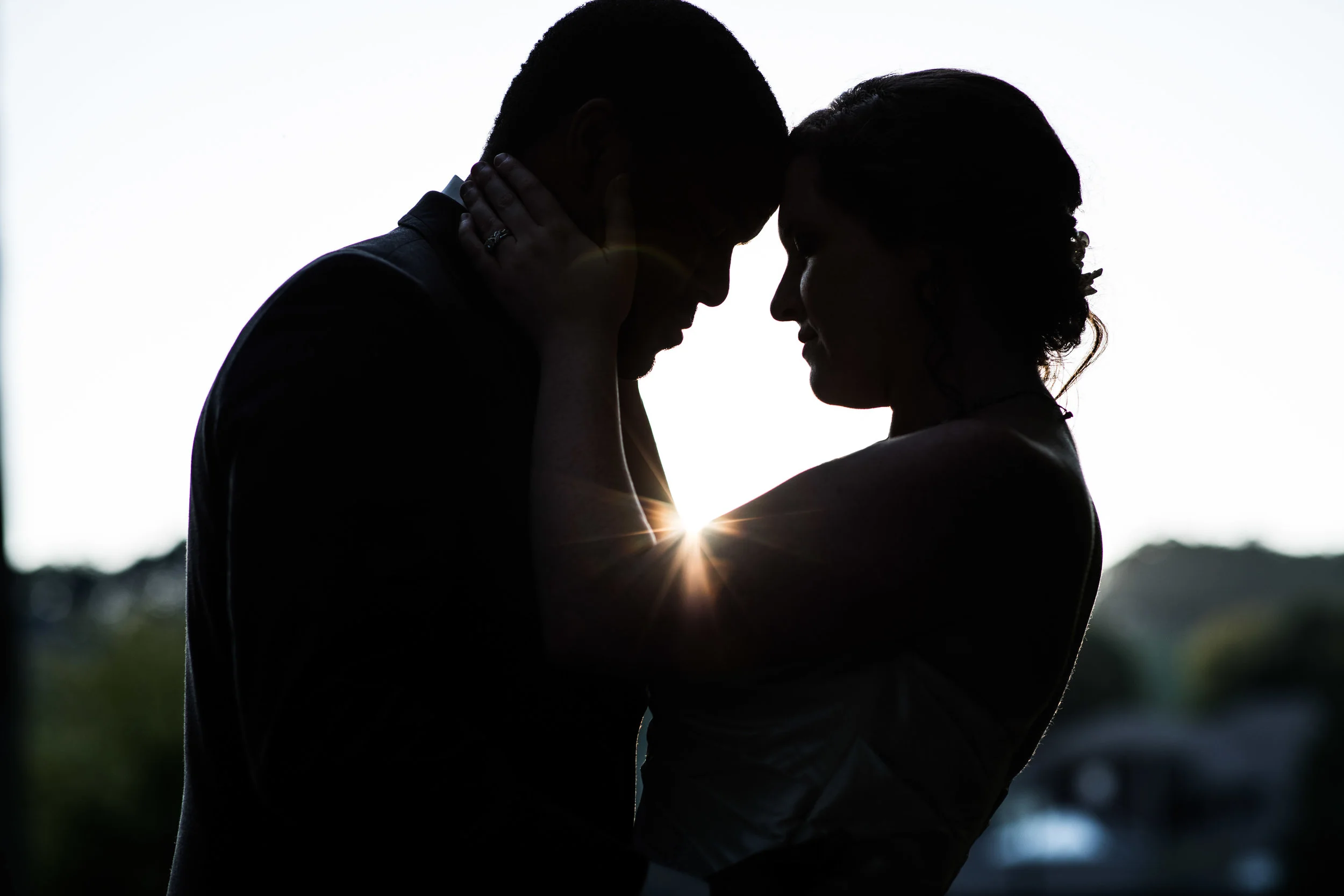 dramatic silhouette of bride and groom holding each other as the setting sun shines through between them