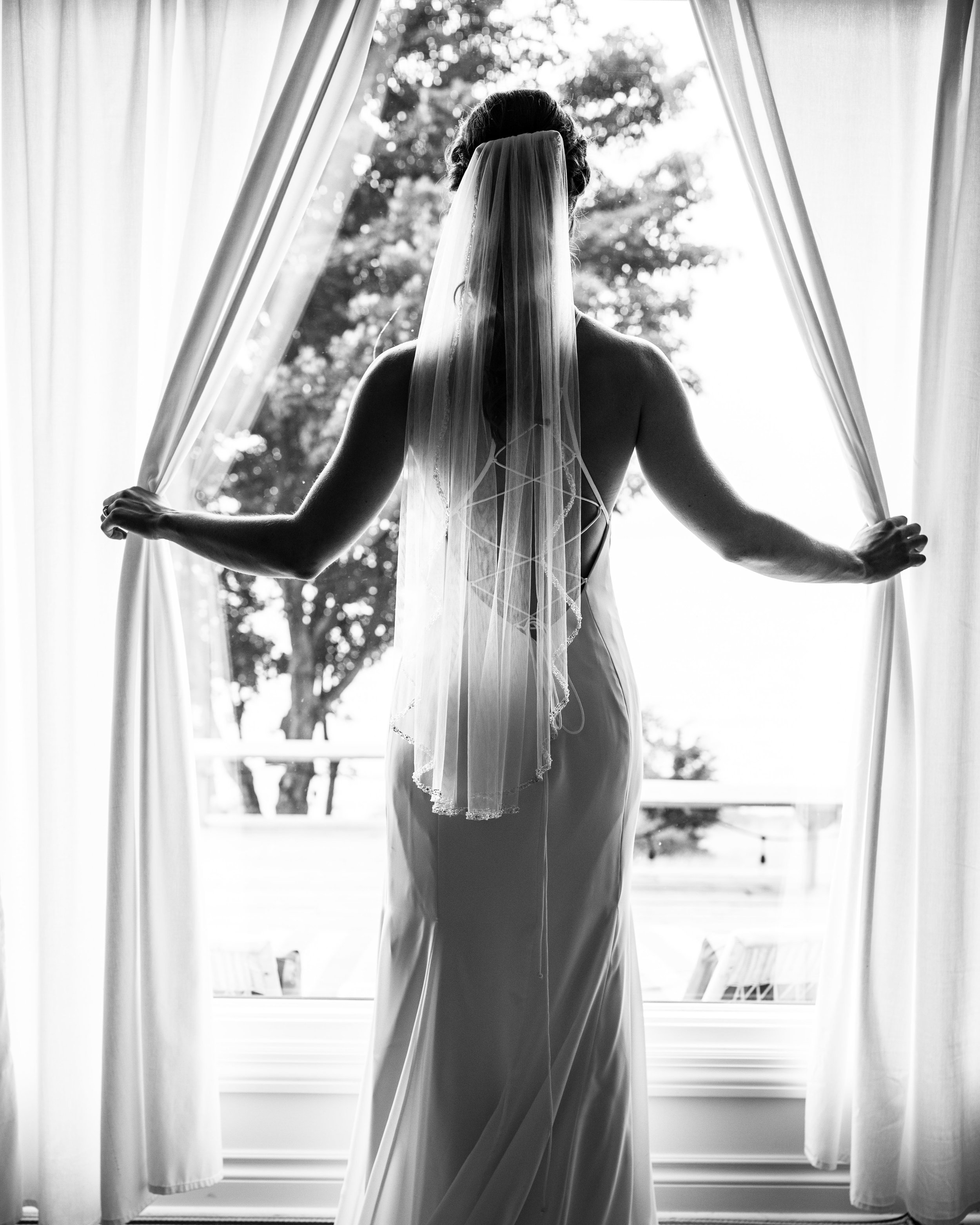  the bride stands in her white dress and veil, opening the curtains to watch the groom arrive 