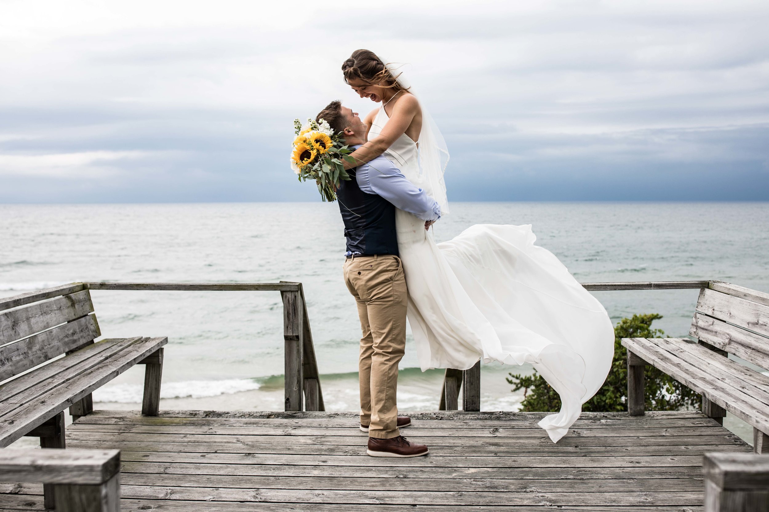  the groom lifts the bride up into the air standing at an overlook to Lake Michigan as her white dress blows in the wind 