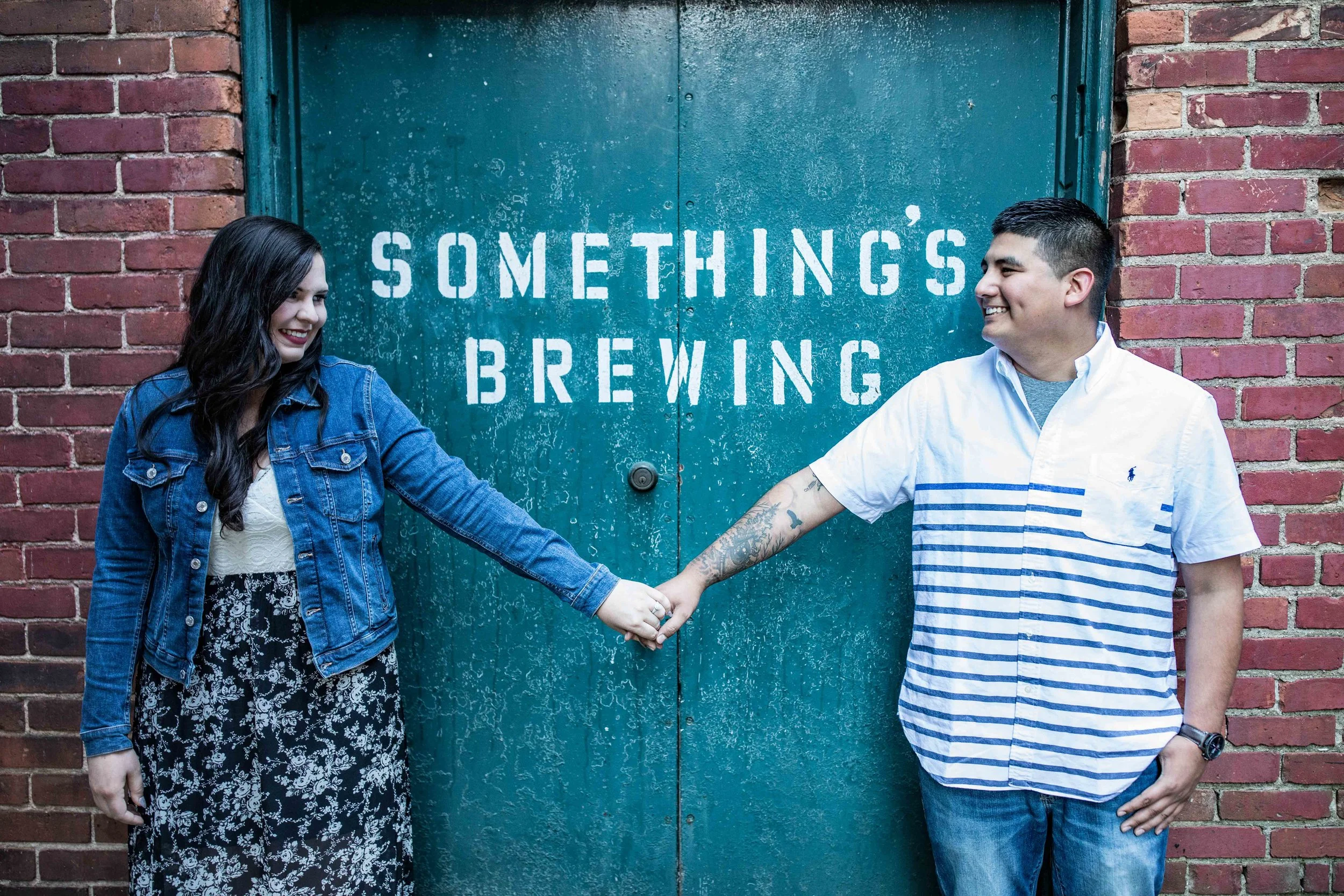 Man and woman hold hands in front of a green double door that says Something’s Brewing