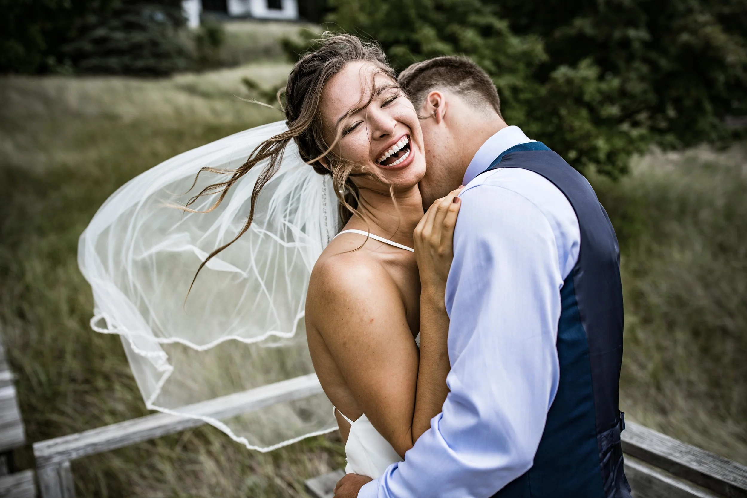 He kisses her neck as she laughs with the wind blowing her hair and veil