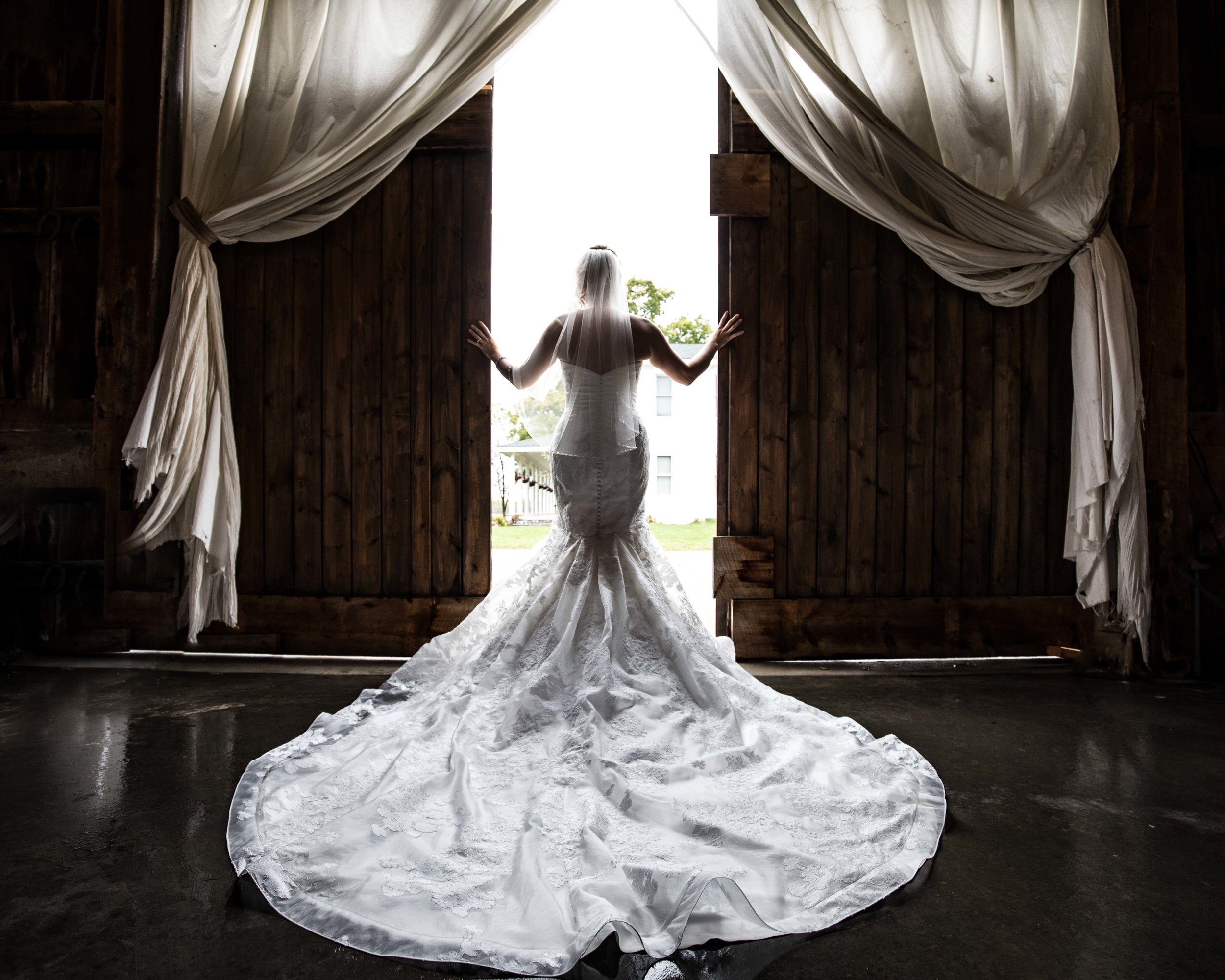  Bride in a white dress with the train spread out as she pushes open the barn doors 