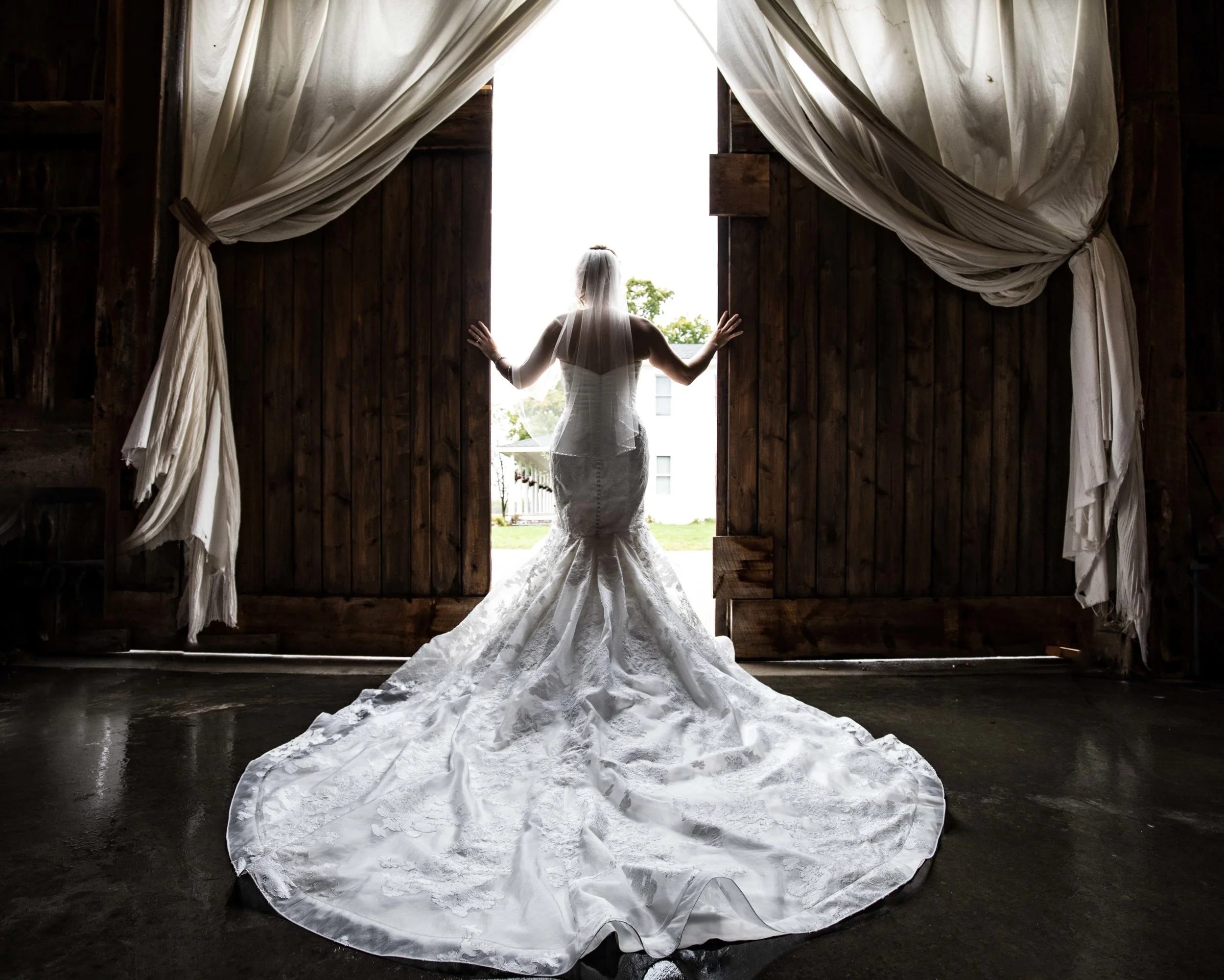 Bride in a white dress with the train spread out as she pushes open the barn doors