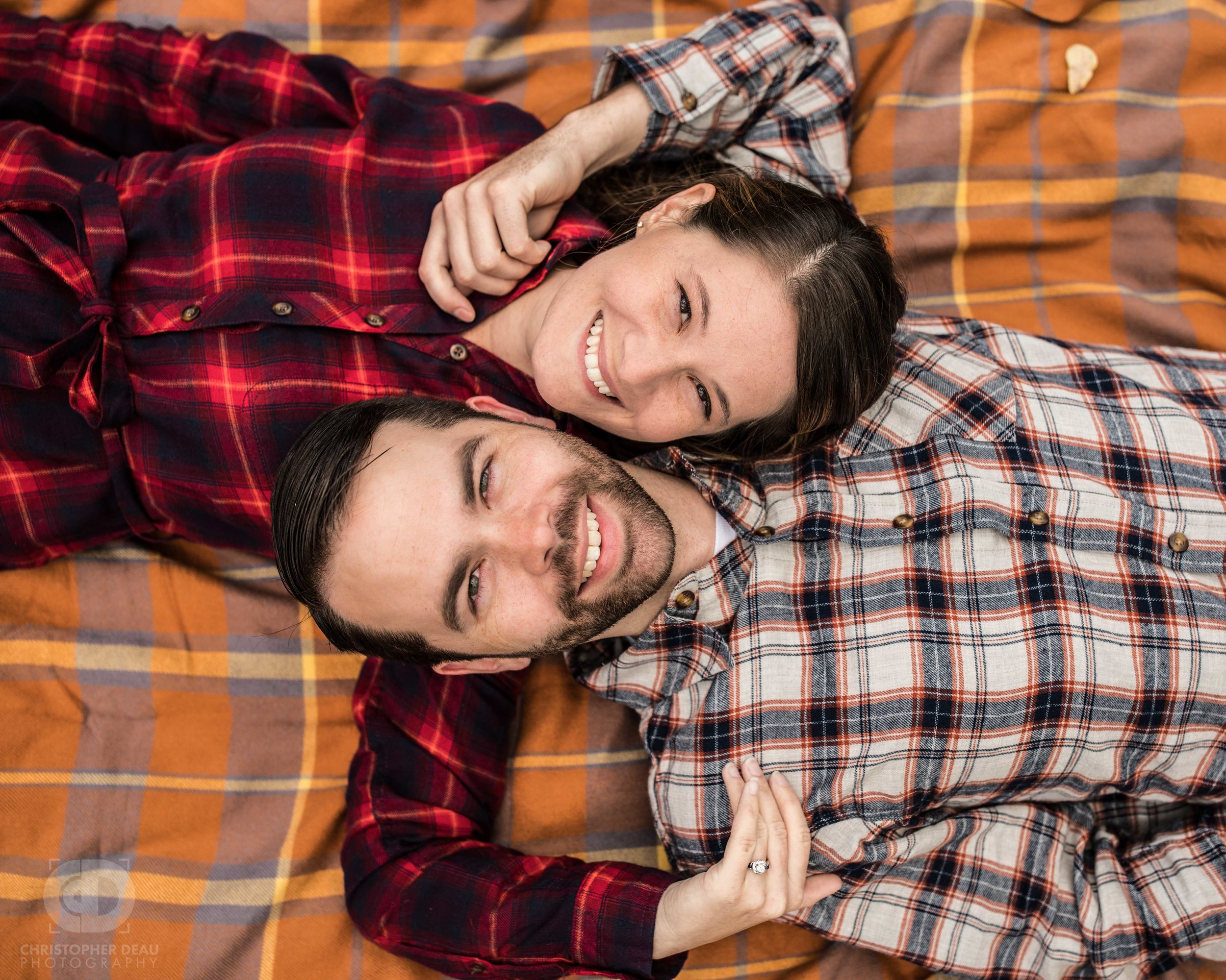  engaged couple laying down on fall blanket, looking up at camera 