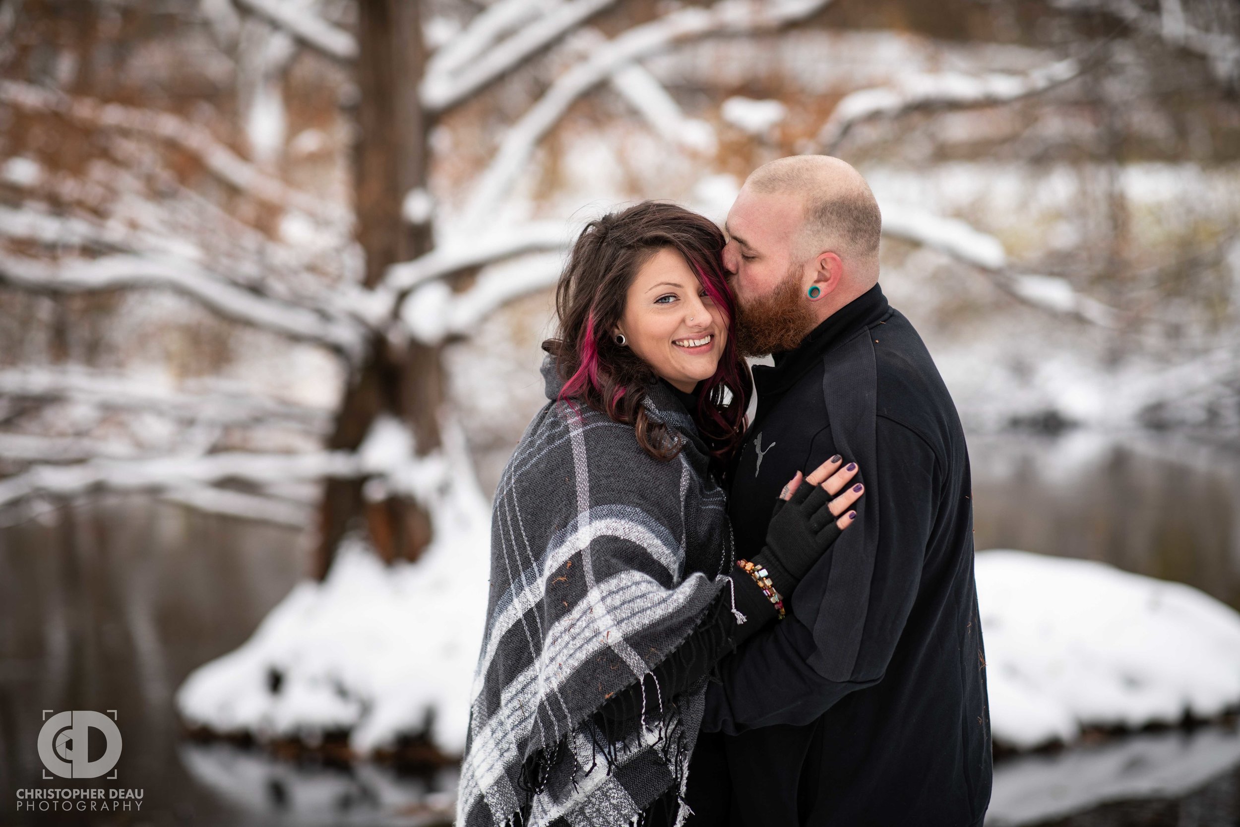  Man kisses a woman’s head as she smiles at the camera with a stream and tree in background 