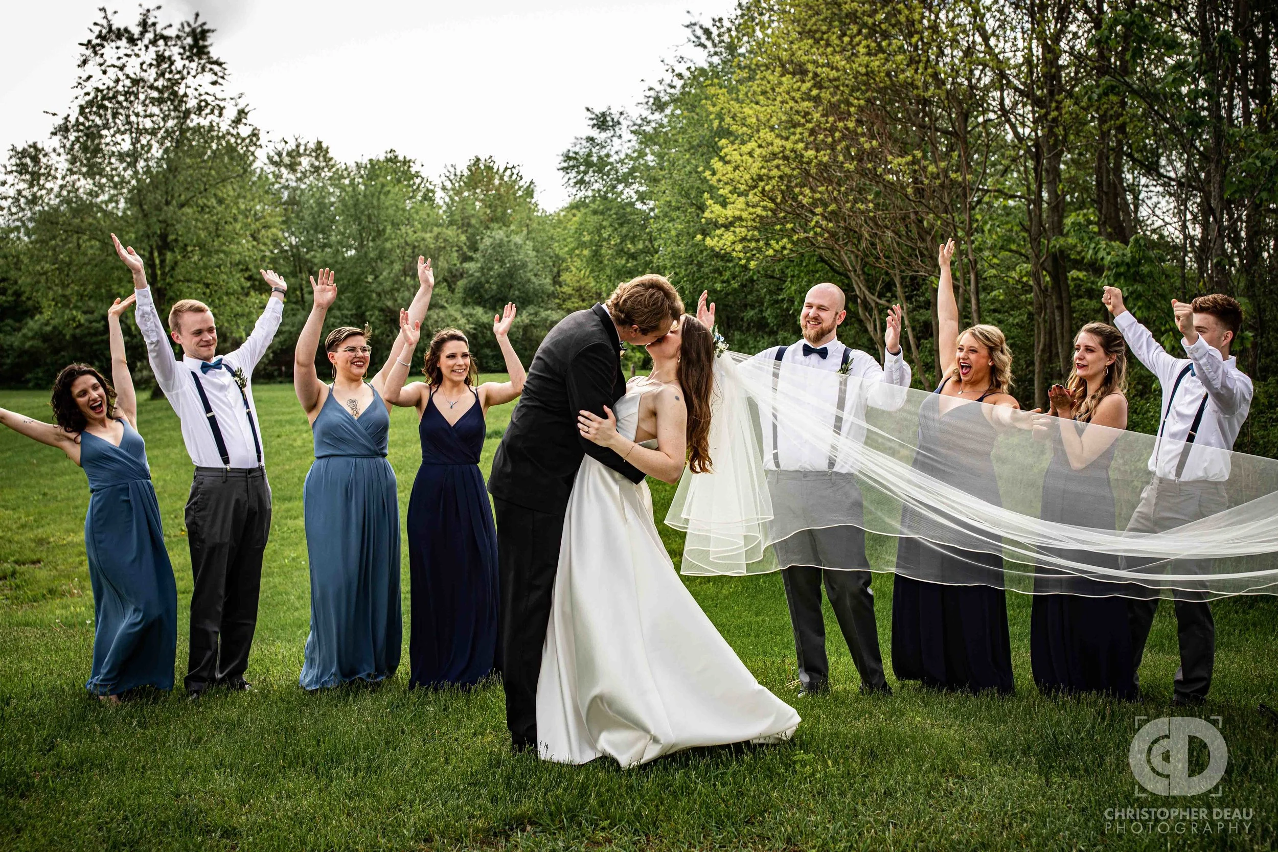 wedding party cheers as bride and groom kiss with long flowing veil