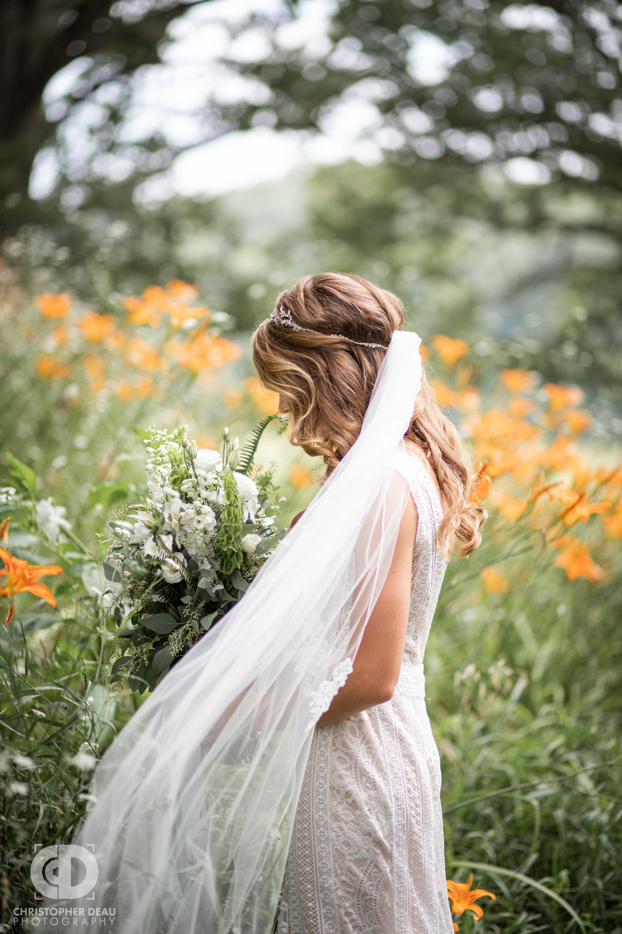  a bride’s veil flows around her as she smells her bouquet while surrounded by flowers 