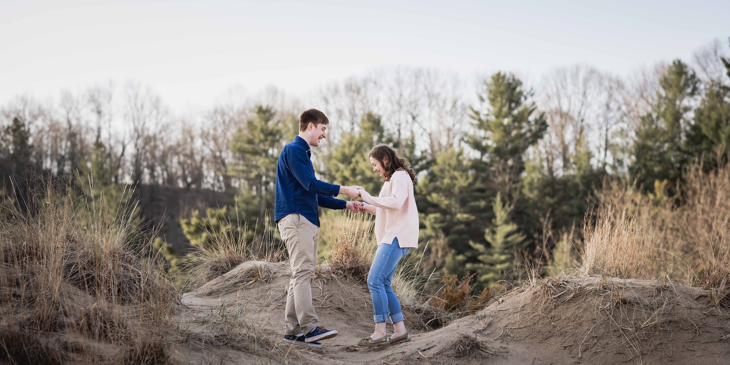  A couple holding hands on a sandy dune with tall green pines in the background 